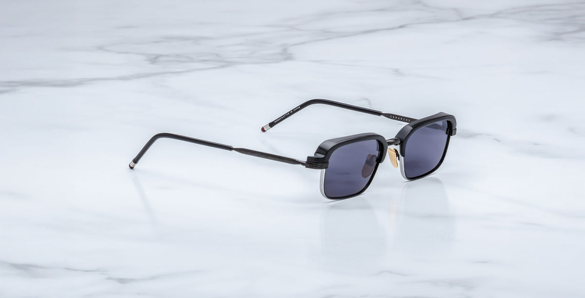 The Jacques Marie Mage Comanche, black square-frame handcrafted sunglasses with dark lenses, rests on a white marble surface.