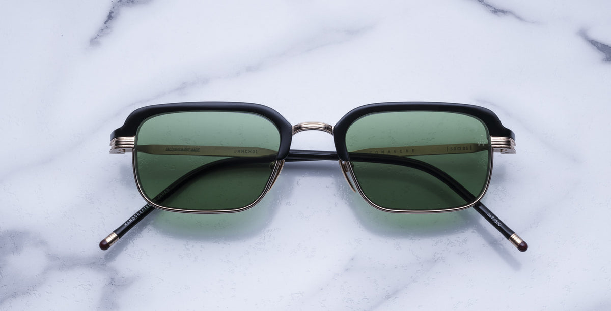The Jacques Marie Mage Comanche sunglasses, featuring square black frames and green lenses, are displayed on a white marble surface.