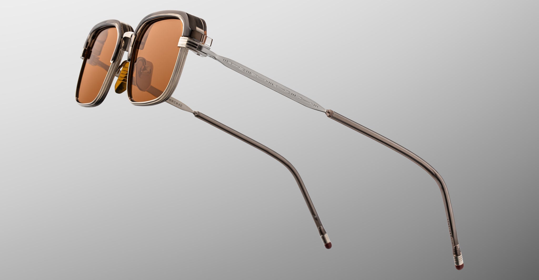 The Jacques Marie Mage Comanche sunglasses feature a square shape, brown-tinted lenses, and slim metallic arms, exemplifying the brand's handcrafted artistry against a neutral backdrop.