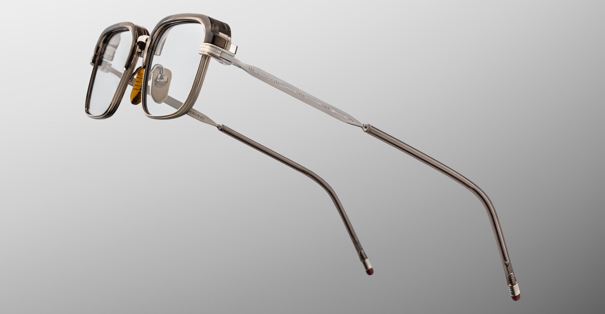 The Jacques Marie Mage Comanche features rectangular clear frames with dark edges and metal temples, shown on a neutral gray background—a limited-edition eyewear piece by Jacques Marie Mage.