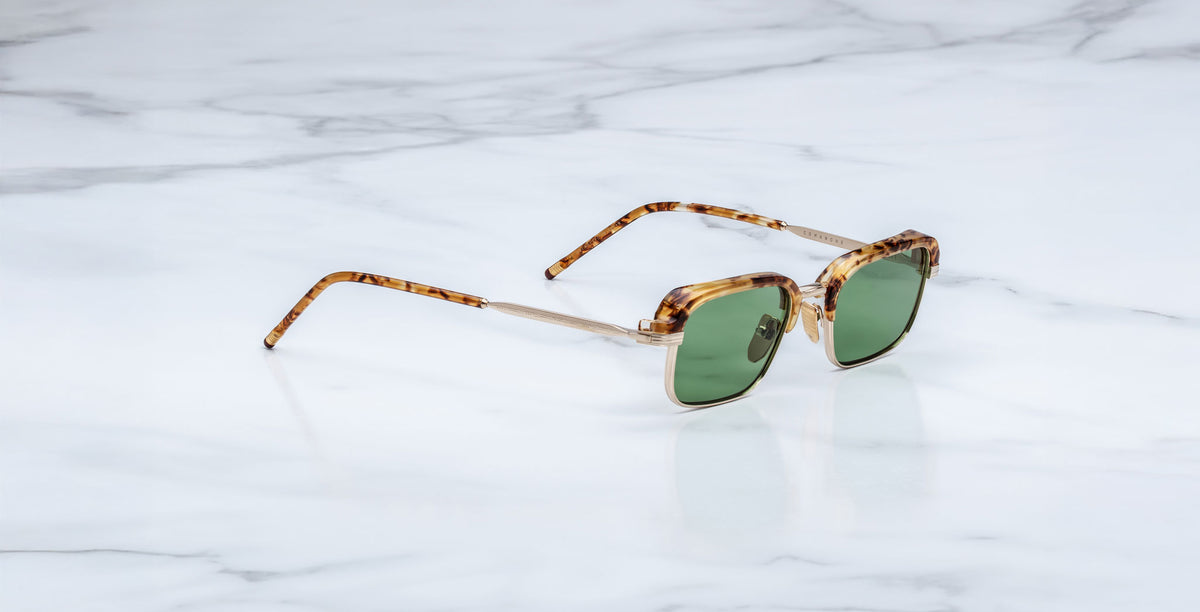 The Jacques Marie Mage Comanche sunglasses, handcrafted with tortoiseshell frames and green lenses, rest on a white marble surface.
