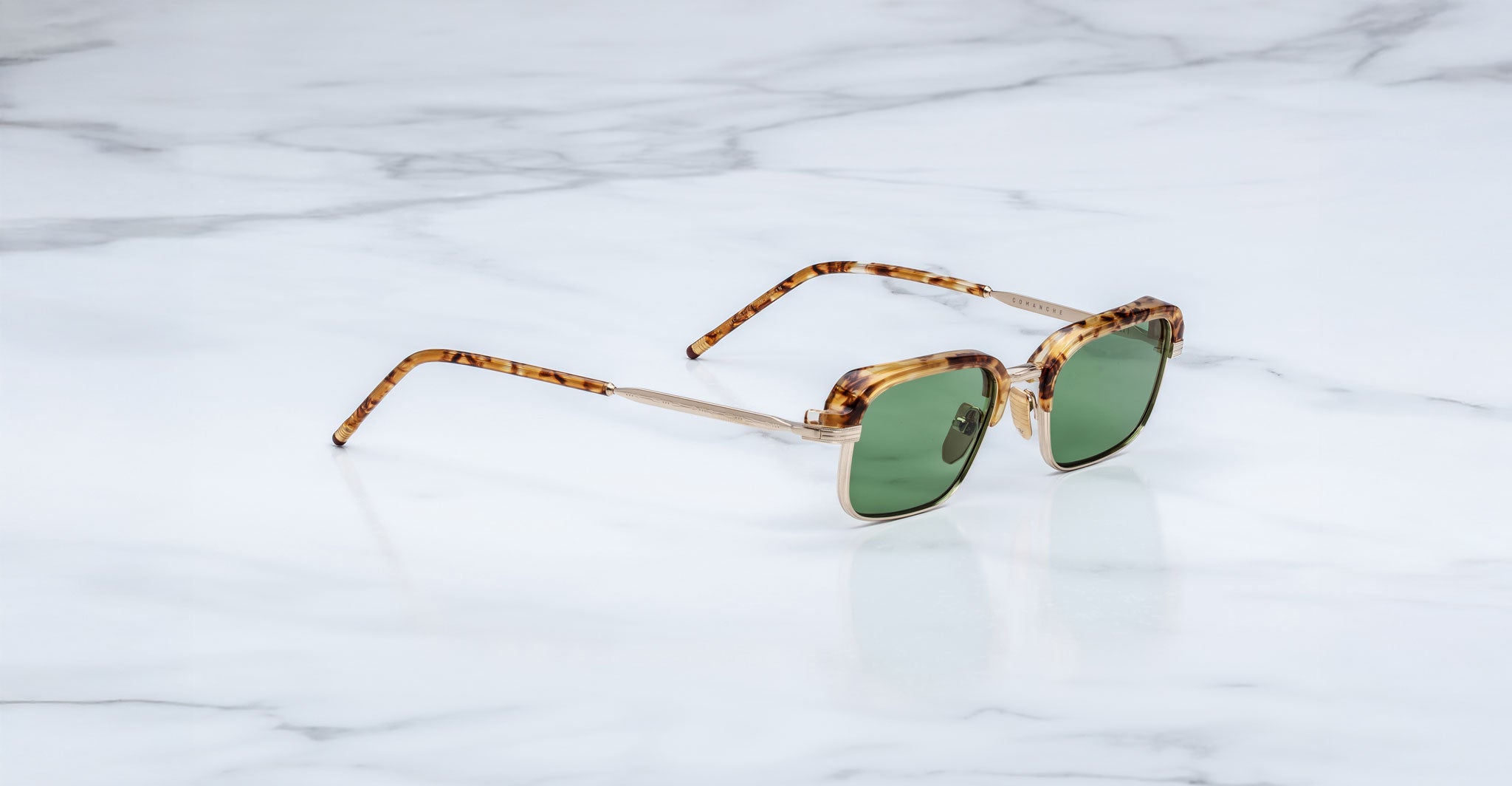 The Jacques Marie Mage Comanche sunglasses, handcrafted with tortoiseshell frames and green lenses, rest on a white marble surface.