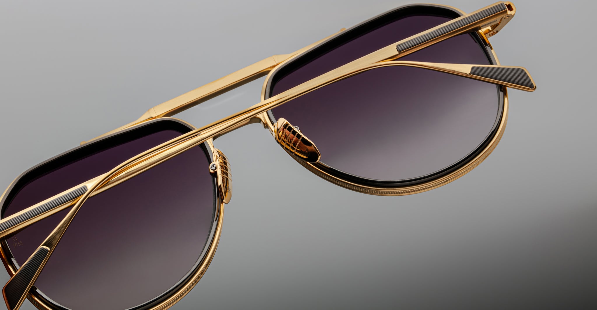 Close-up view of Jacques Marie Mage Commander gold-framed sunglasses with dark gradient lenses on a gray background, highlighting the bridge, nose pads, and temple details.