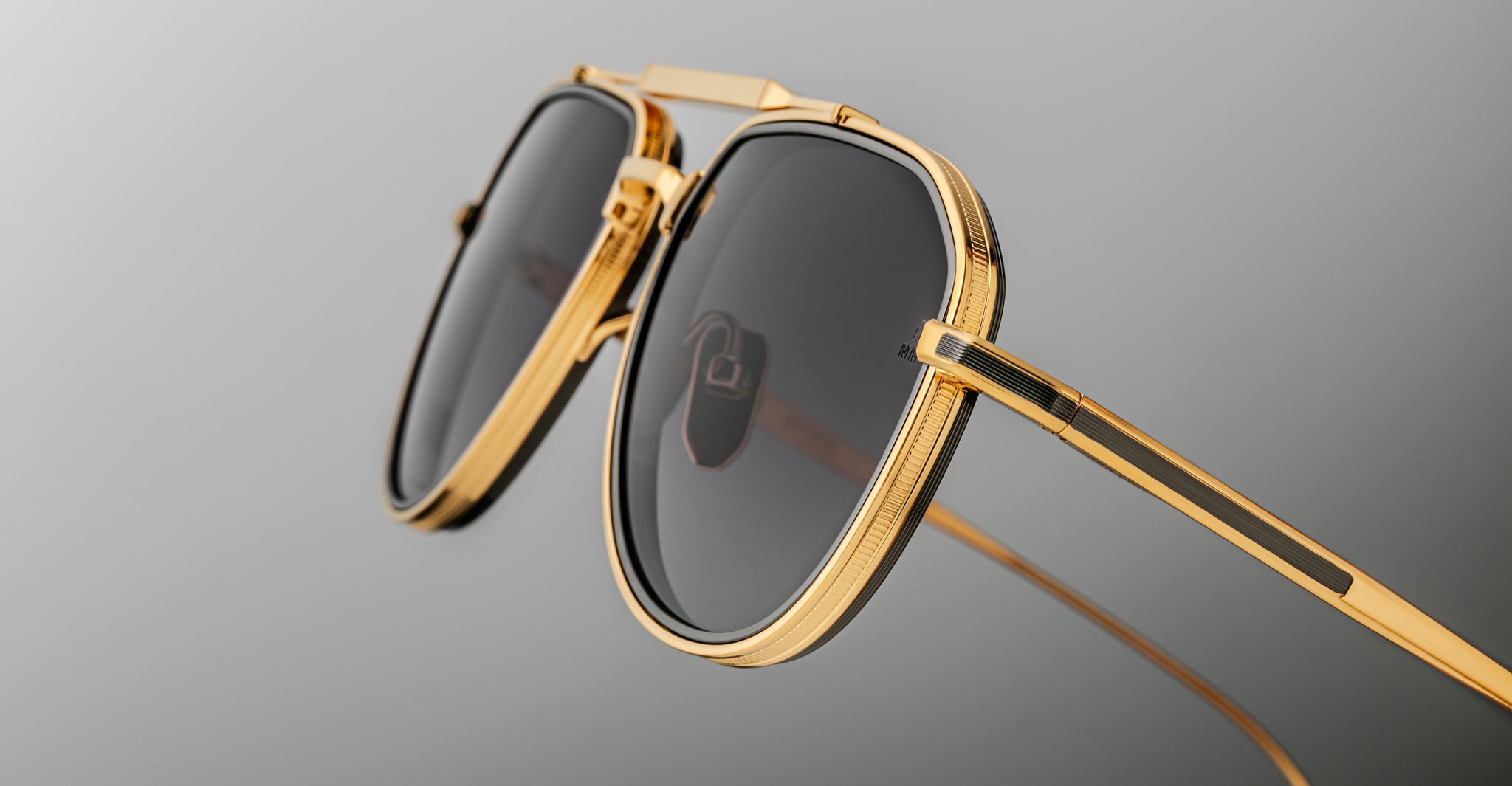 Close-up of Jacques Marie Mage Commander sunglasses with gold frames and dark lenses on a grey background, showcasing their detailed construction and reflective finish.