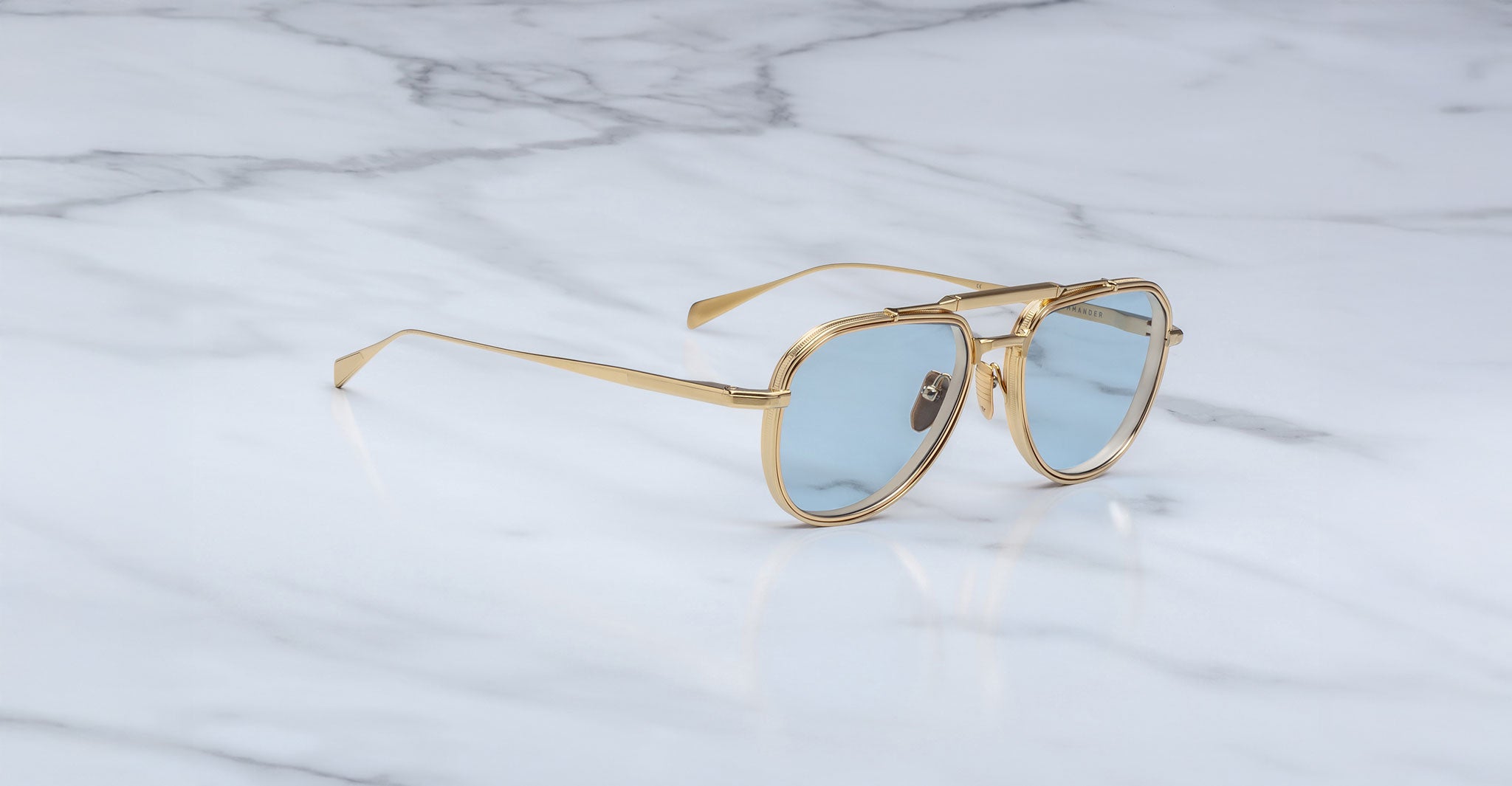 The Jacques Marie Mage Commander gold-framed aviator sunglasses with light blue lenses rest on a white marble surface adorned with gray veining.