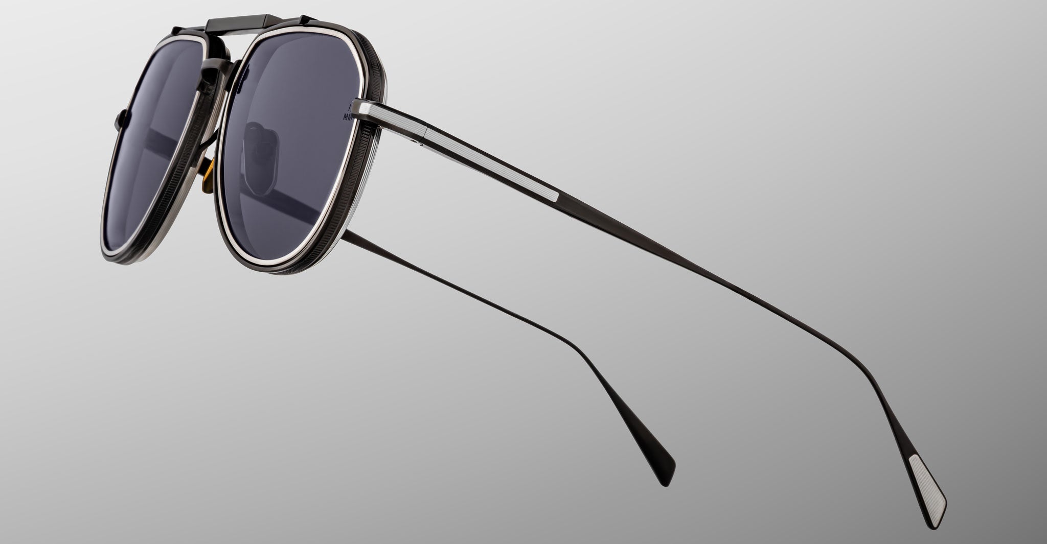 Close-up of Jacques Marie Mage Commander sunglasses with dark lenses, thin black metal frames, and a double bridge design, displayed against a plain gradient gray background.