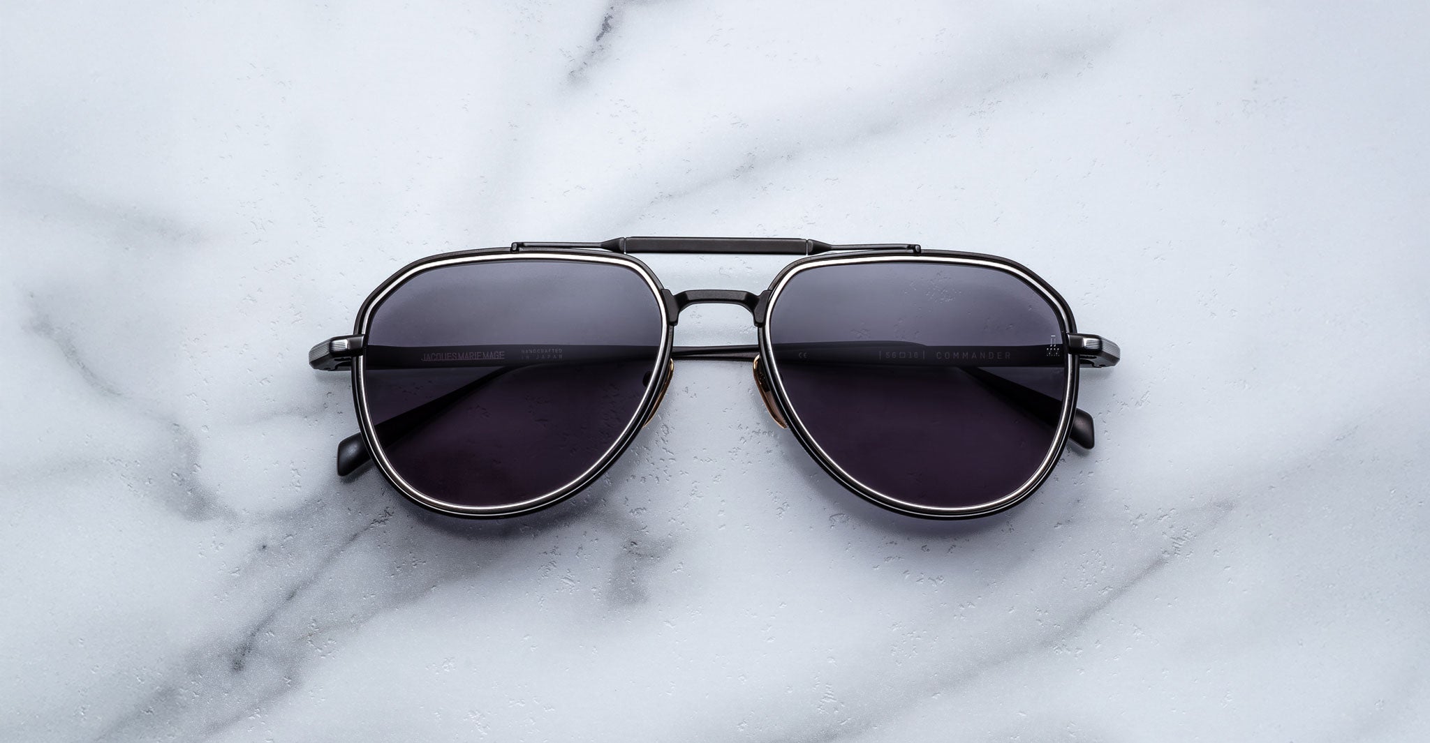 A pair of Jacques Marie Mage Commander black aviator sunglasses with dark lenses and silver rims rests on a white marble surface.
