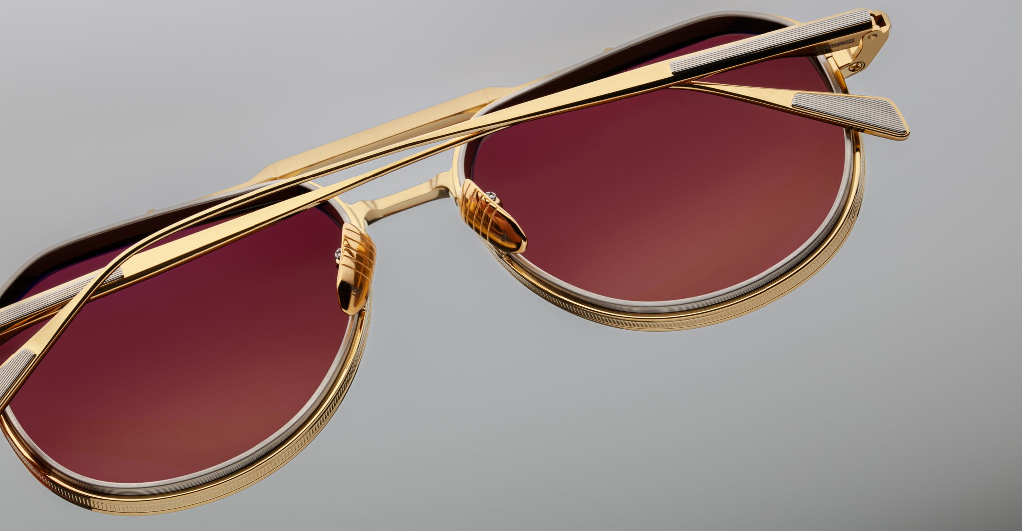 Close-up of Jacques Marie Mage Commander sunglasses with gold frames, red-tinted lenses, and a geometric design set against a neutral gray background.