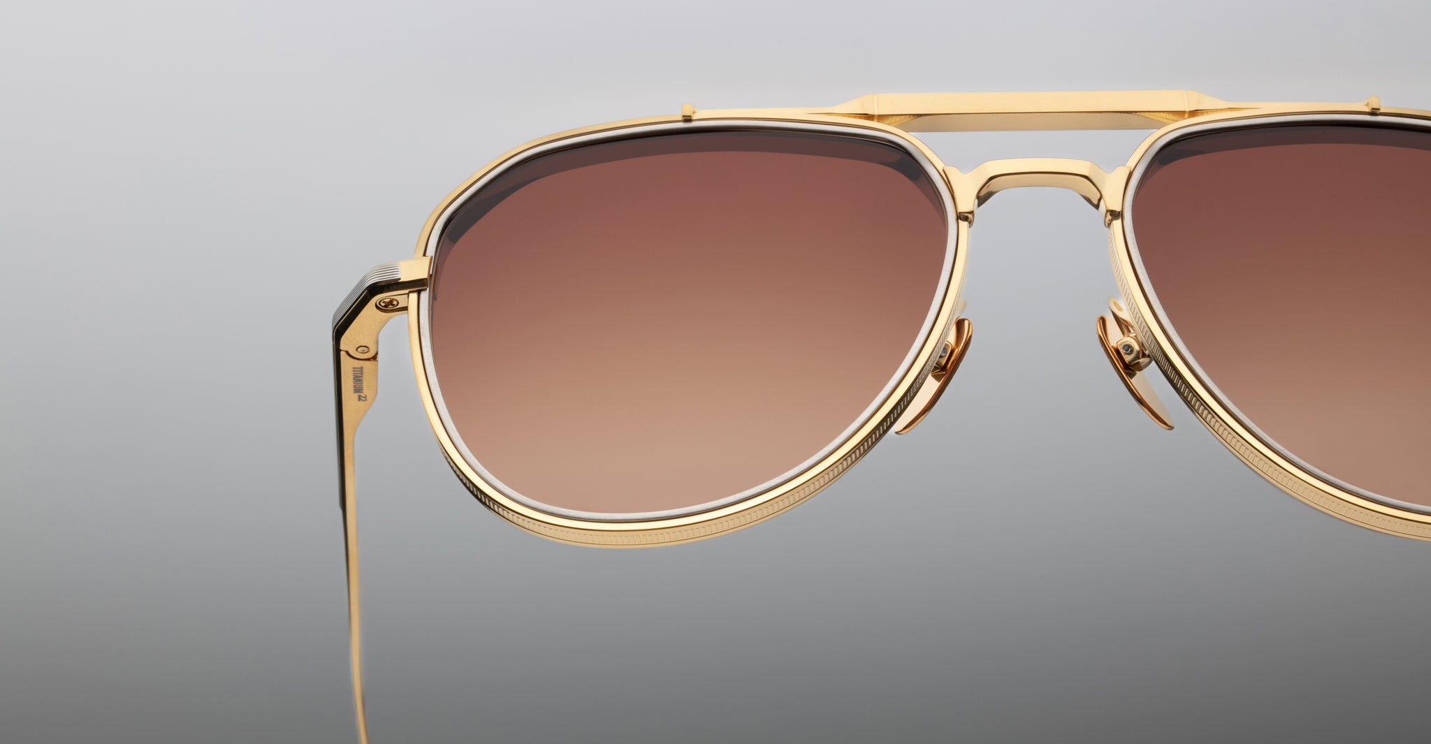 Close-up of the right side of Jacques Marie Mage Commander gold-framed aviator sunglasses with brown gradient lenses against a plain, light gray background.