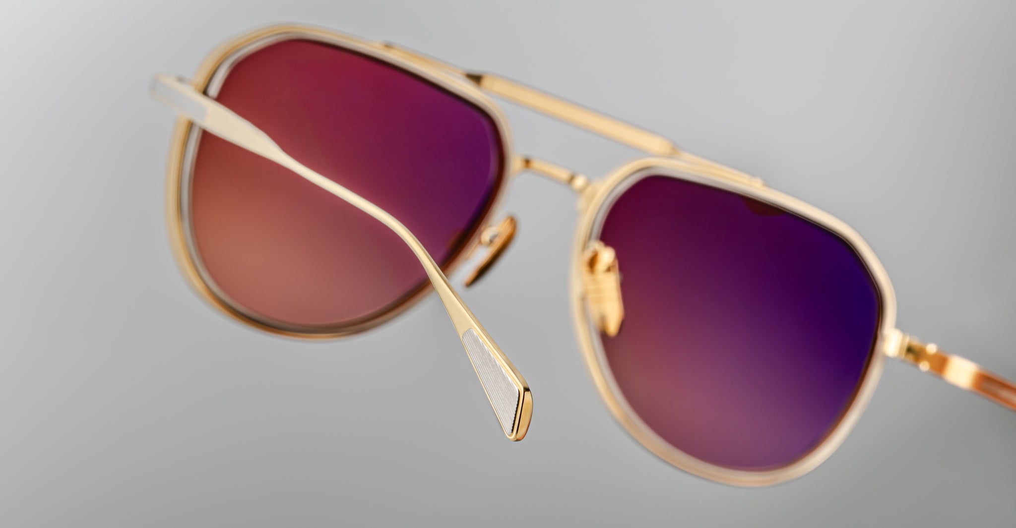 A close-up of Jacques Marie Mage Commander gold-framed aviator sunglasses with purple-tinted lenses, positioned at an angle against a plain light grey background.