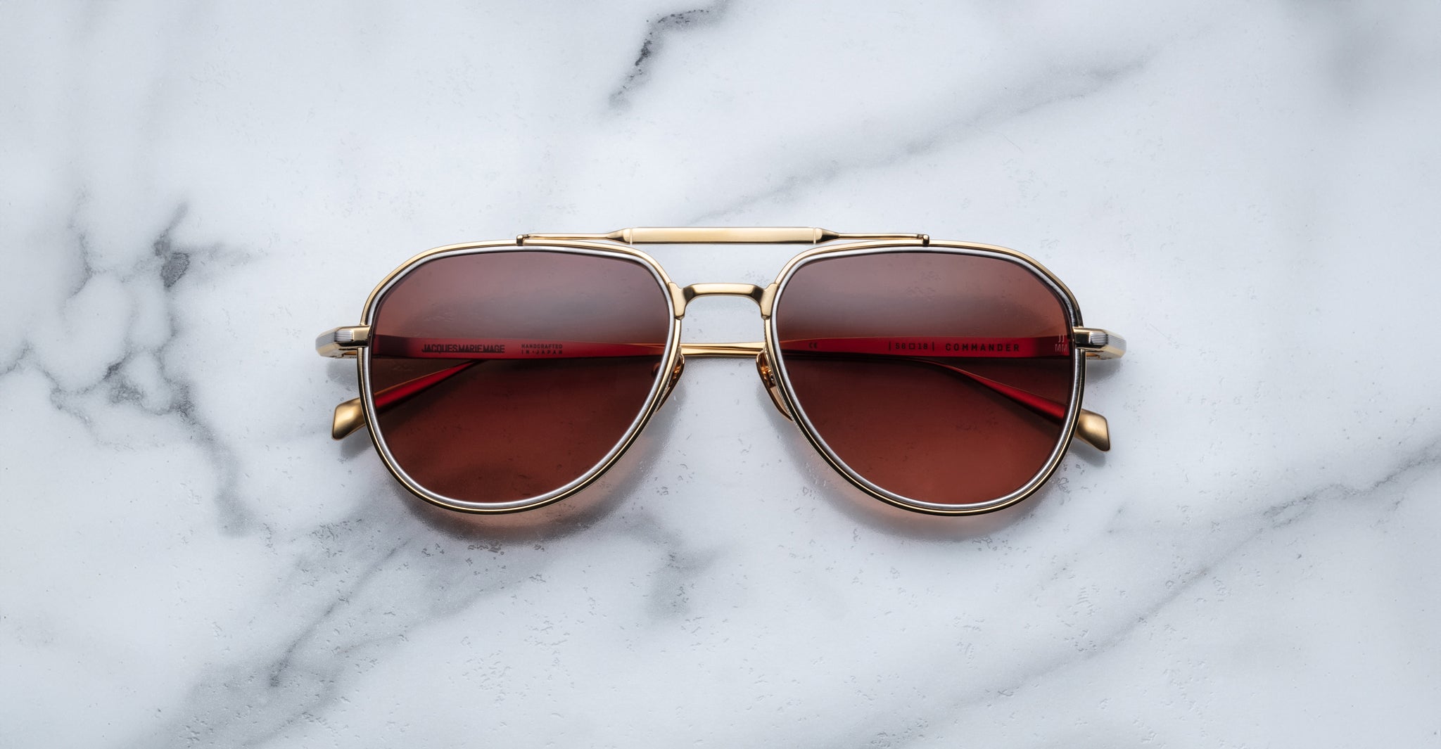 A pair of Jacques Marie Mage Commander gold-framed aviator sunglasses with red-tinted lenses rests on a white marble surface.