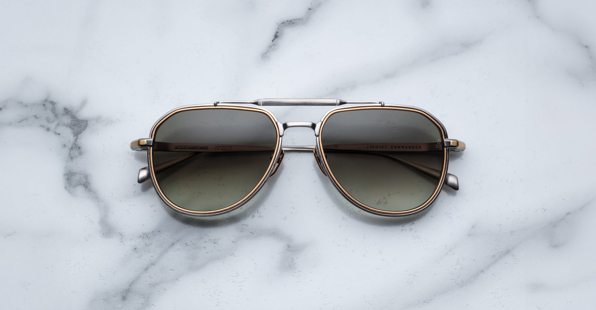A pair of Jacques Marie Mage Commander aviator sunglasses with metal frames and tinted lenses rests on a white marble surface with gray veining.