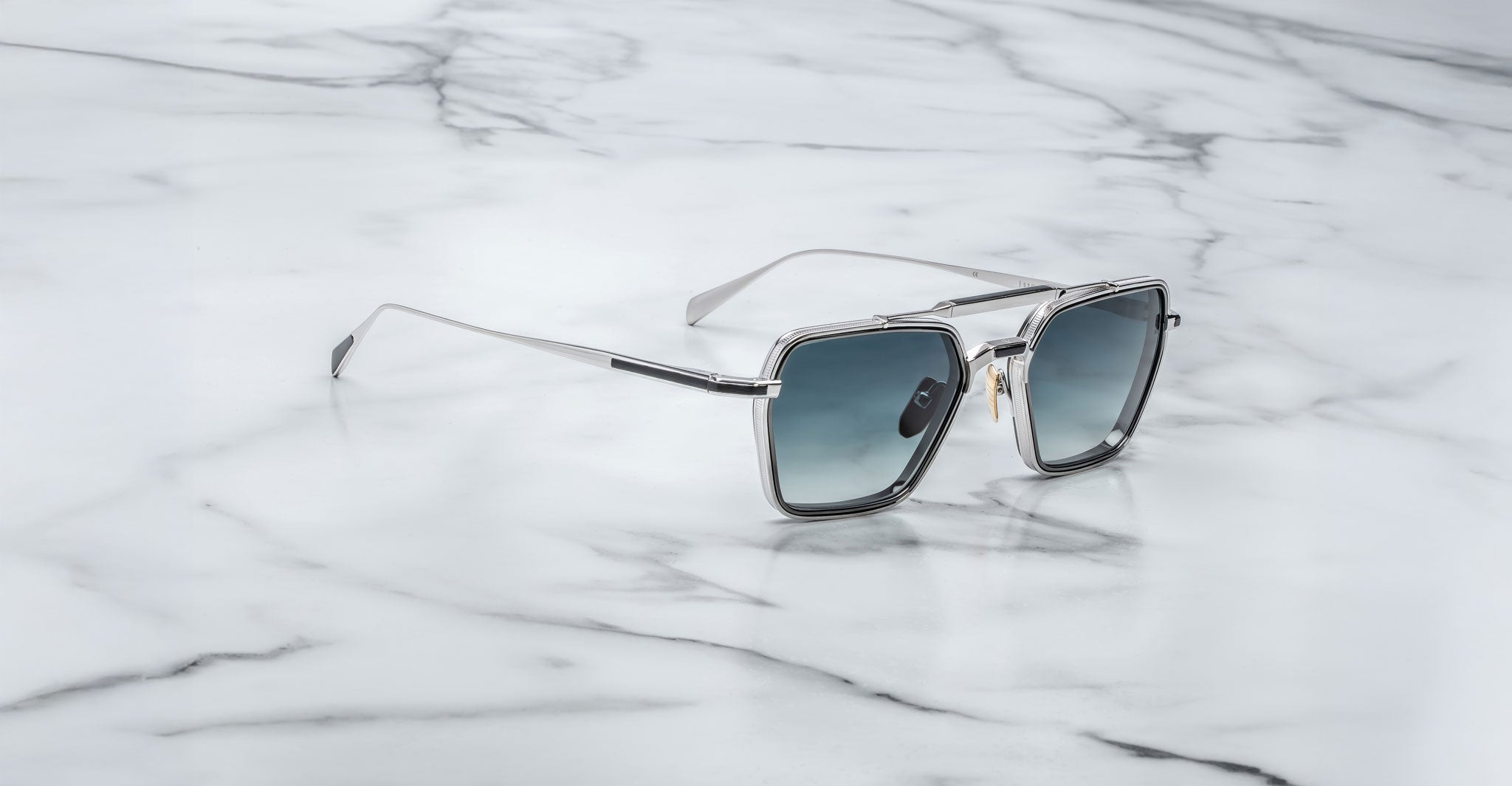 Jacques Marie Mage Commodore square sunglasses with silver metal frames and blue tinted lenses are displayed on a white marble surface with gray veining. The minimalistic setting highlights their modern, luxurious design.