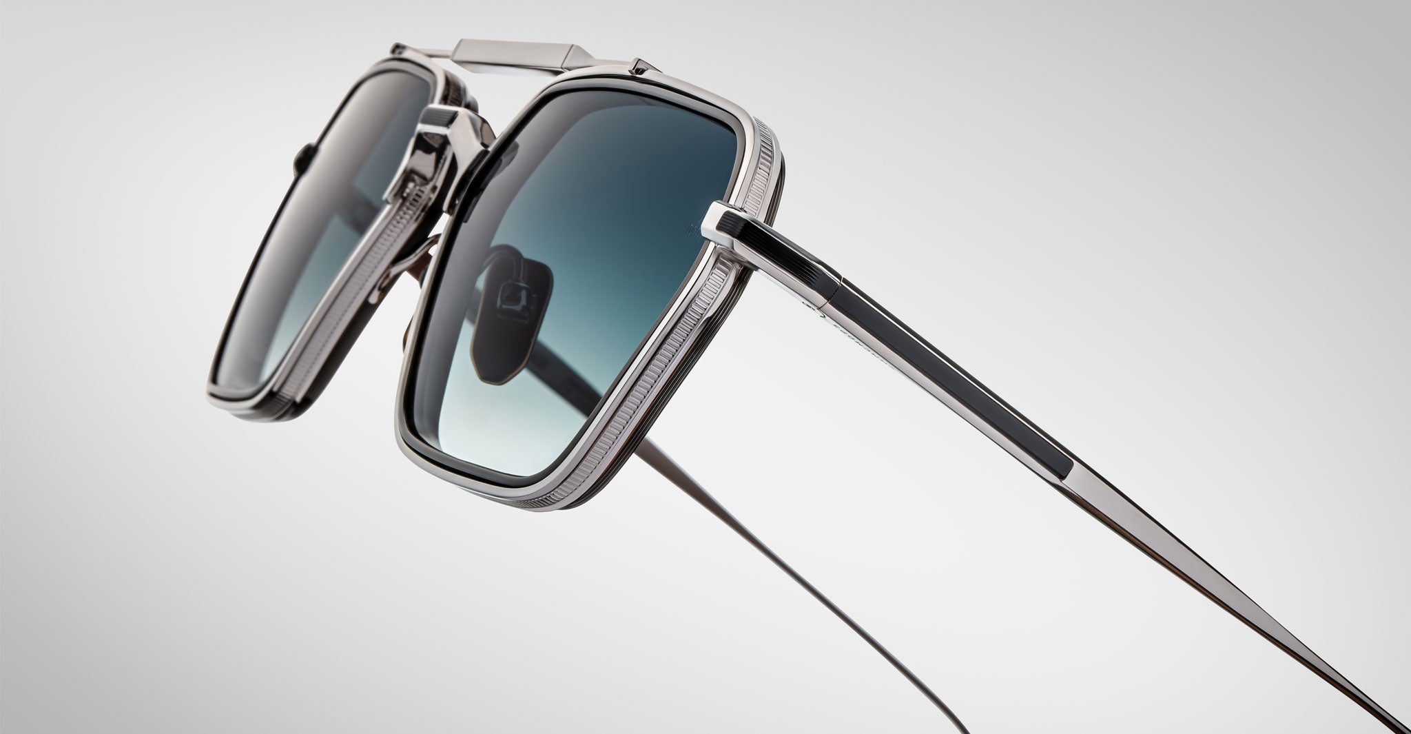 Close-up of Jacques Marie Mage Commodore sunglasses with square silver metal frames and mirrored lenses, displayed at an angle on a smooth, light gray background.