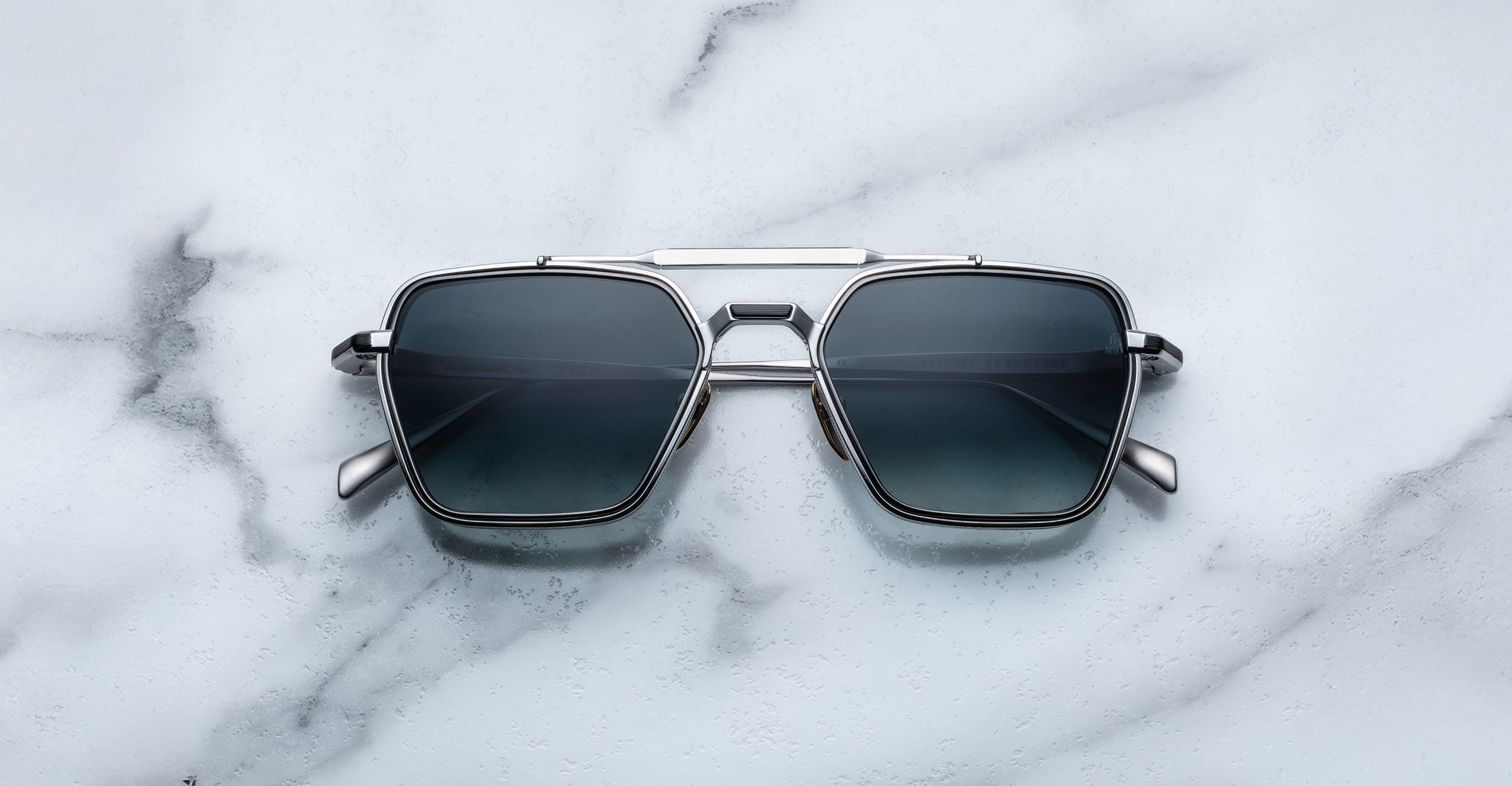 The Jacques Marie Mage Commodore features a modern rectangular design with dark lenses and a silver metal frame, showcased on a white marble surface.
