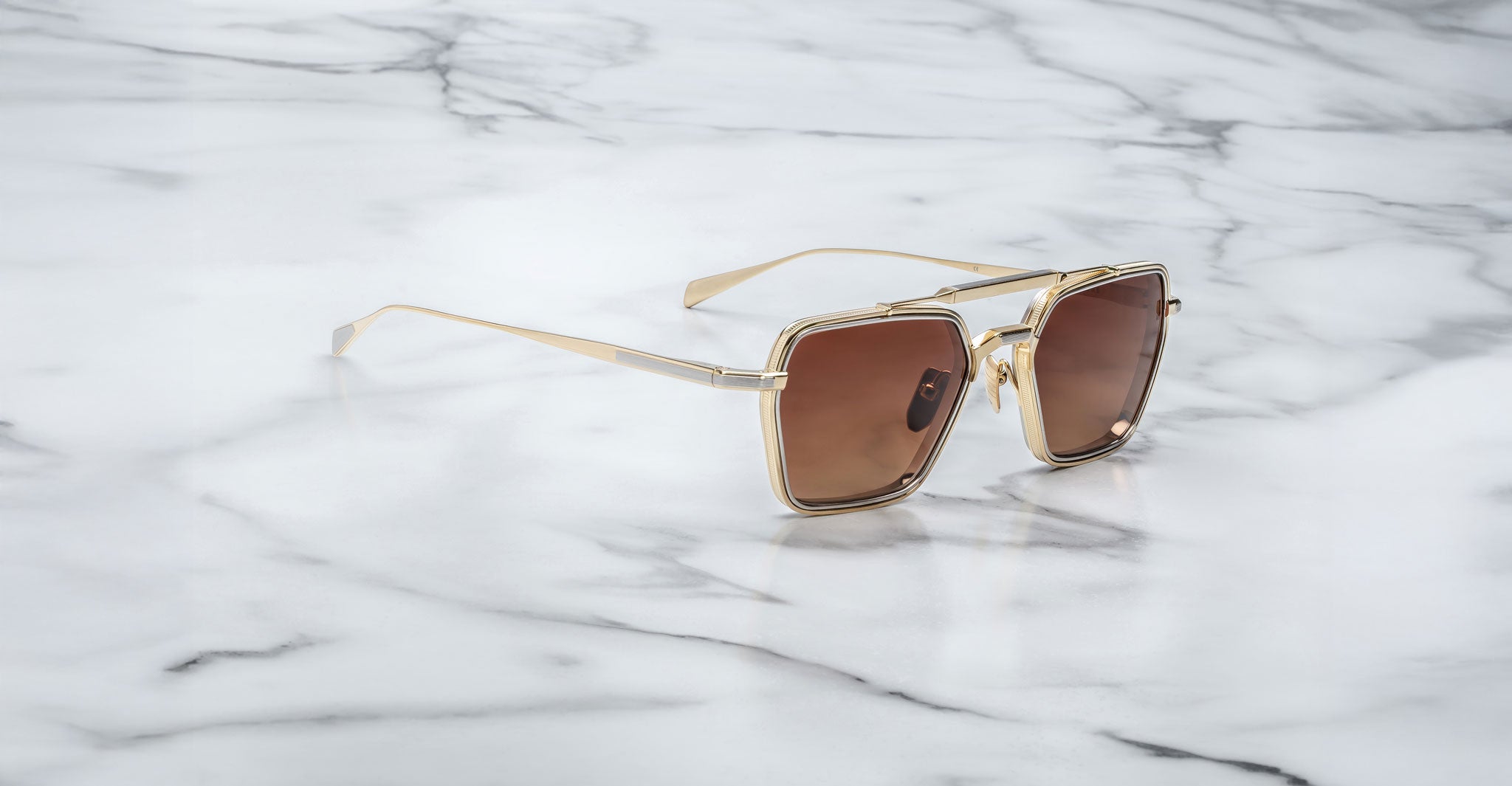 Jacques Marie Mage Commodore gold-framed, square sunglasses with brown lenses placed on a white marble surface featuring grey veining.