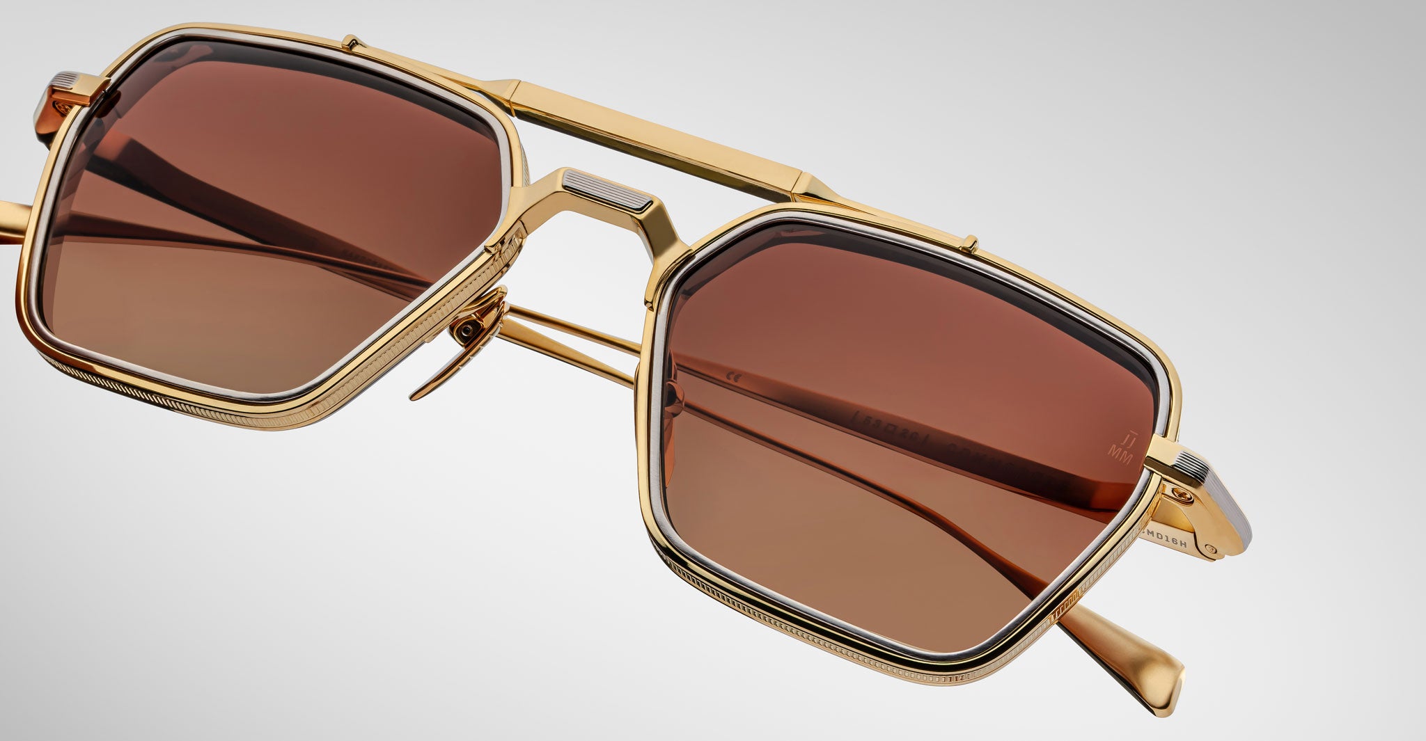 Jacques Marie Mage Commodore sunglasses with gold geometric frames and brown-tinted lenses rest on a light gray background. Bold, angular lines and a sturdy bridge showcase their modern, luxurious design.