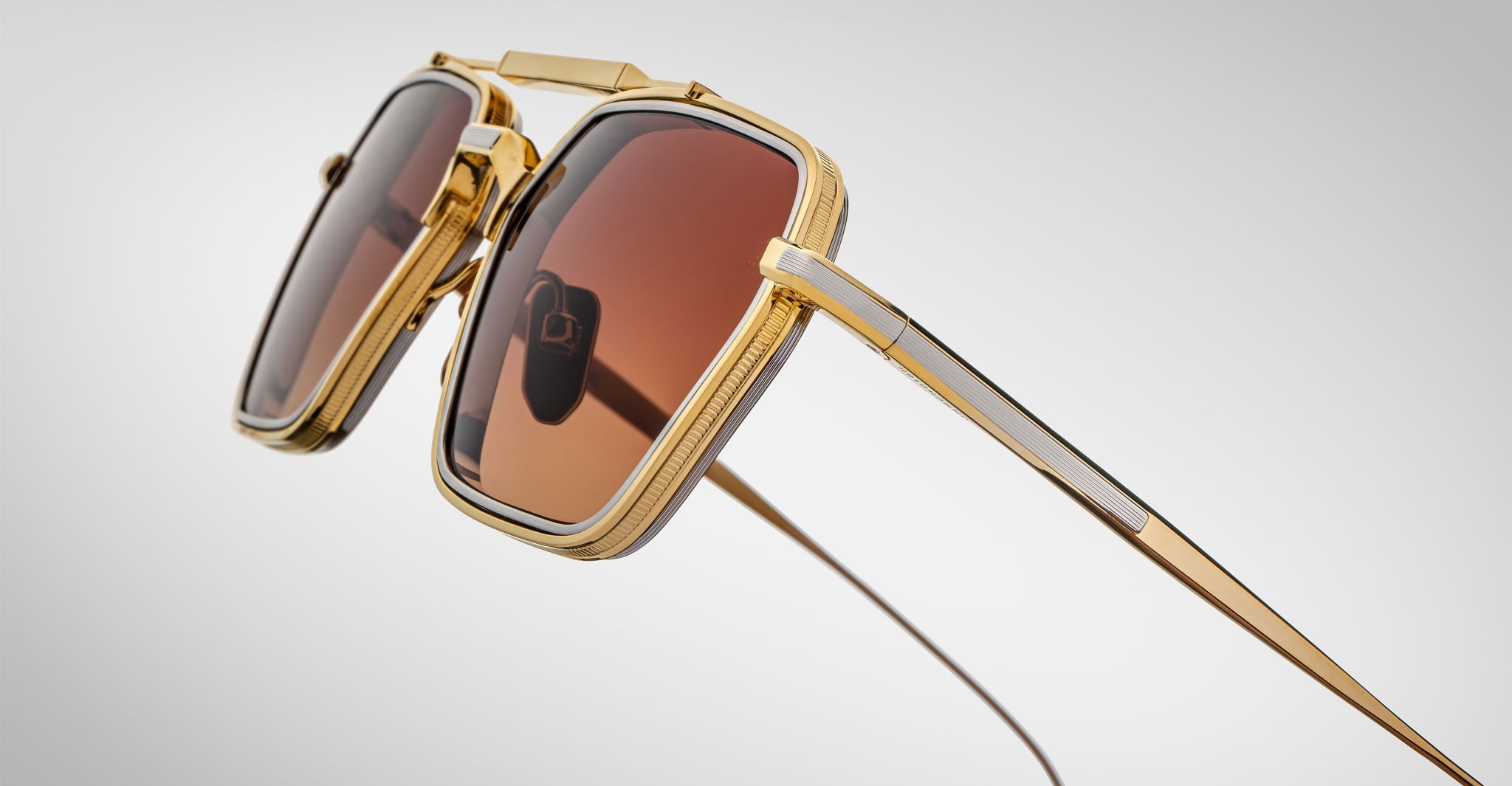 A close-up of Jacques Marie Mage Commodore sunglasses showcases stylish gold frames with brown-tinted square lenses and intricate detailing on the frame and temples, set against a light gray gradient background.