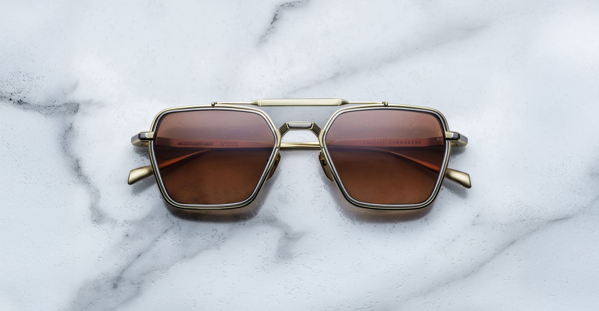 A pair of Jacques Marie Mage Commodore sunglasses with gold square frames and brown lenses rests on a white marble surface.