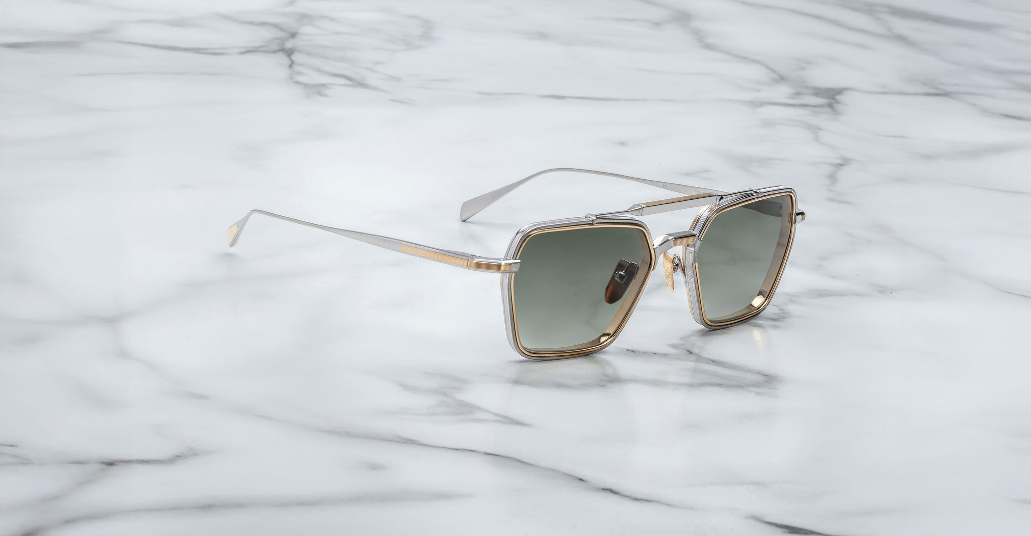 Jacques Marie Mage Commodore sunglasses with gold square frames and dark lenses sit on a white marble surface with gray veining.