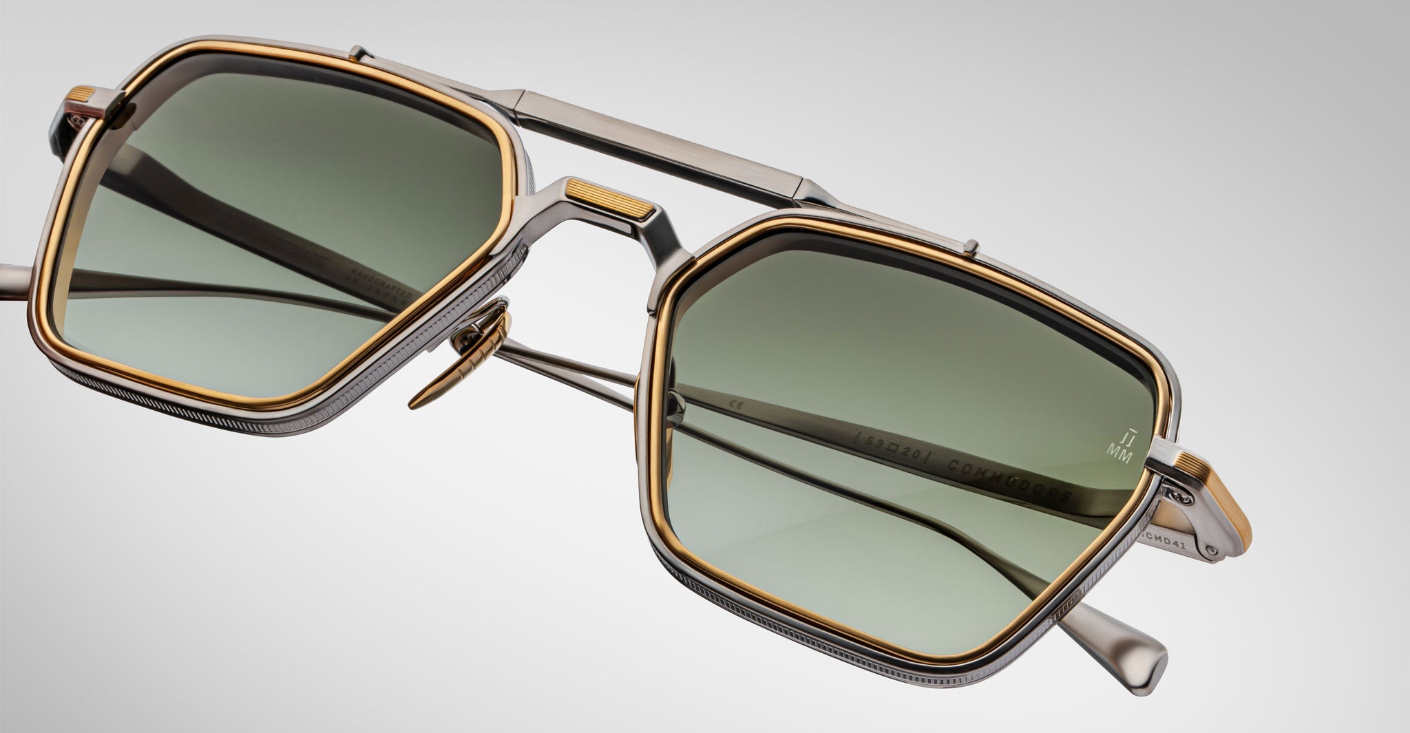 Close-up of Jacques Marie Mage Commodore hexagonal sunglasses featuring gold and silver metal frames, green tinted lenses, and a double bridge design, set against a light gray background.