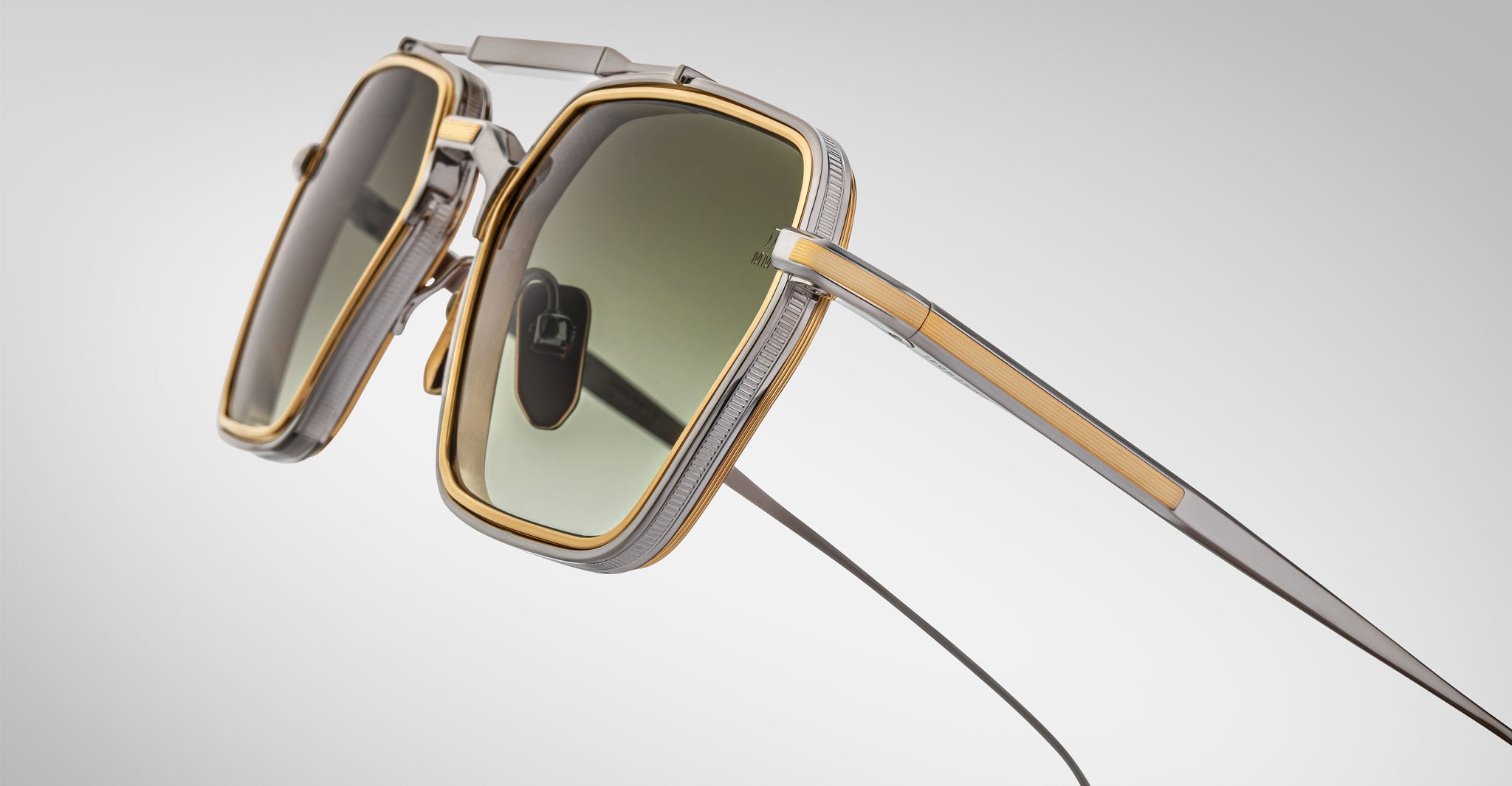Close-up of Jacques Marie Mage Commodore sunglasses featuring a metallic silver and gold rectangular frame, green-tinted lenses, and uniquely designed thin temples against a light gray gradient background.