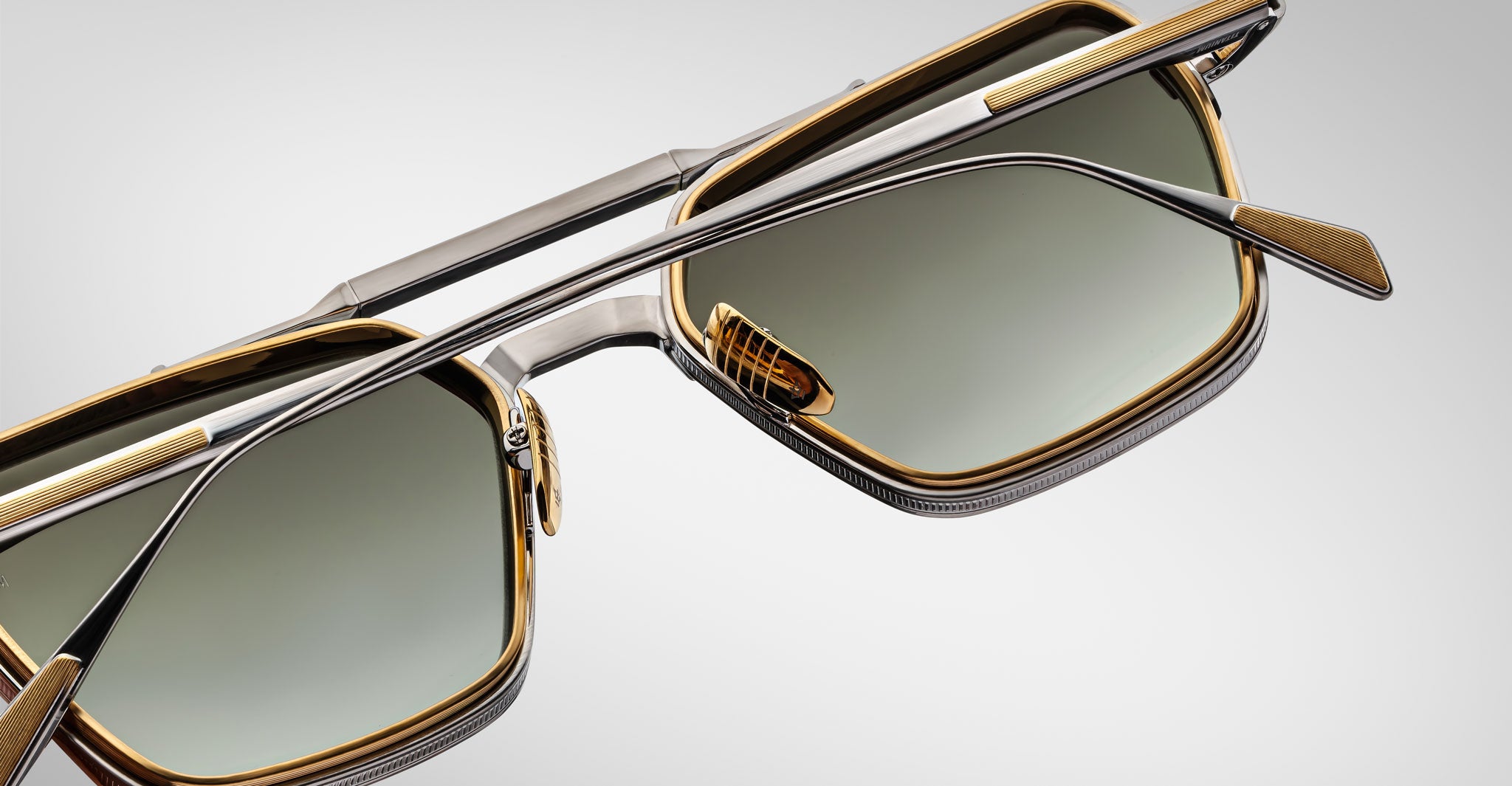 Close-up of Jacques Marie Mage Commodore sunglasses featuring square green-tinted lenses and a gold and silver metallic frame, set against a light gray background.