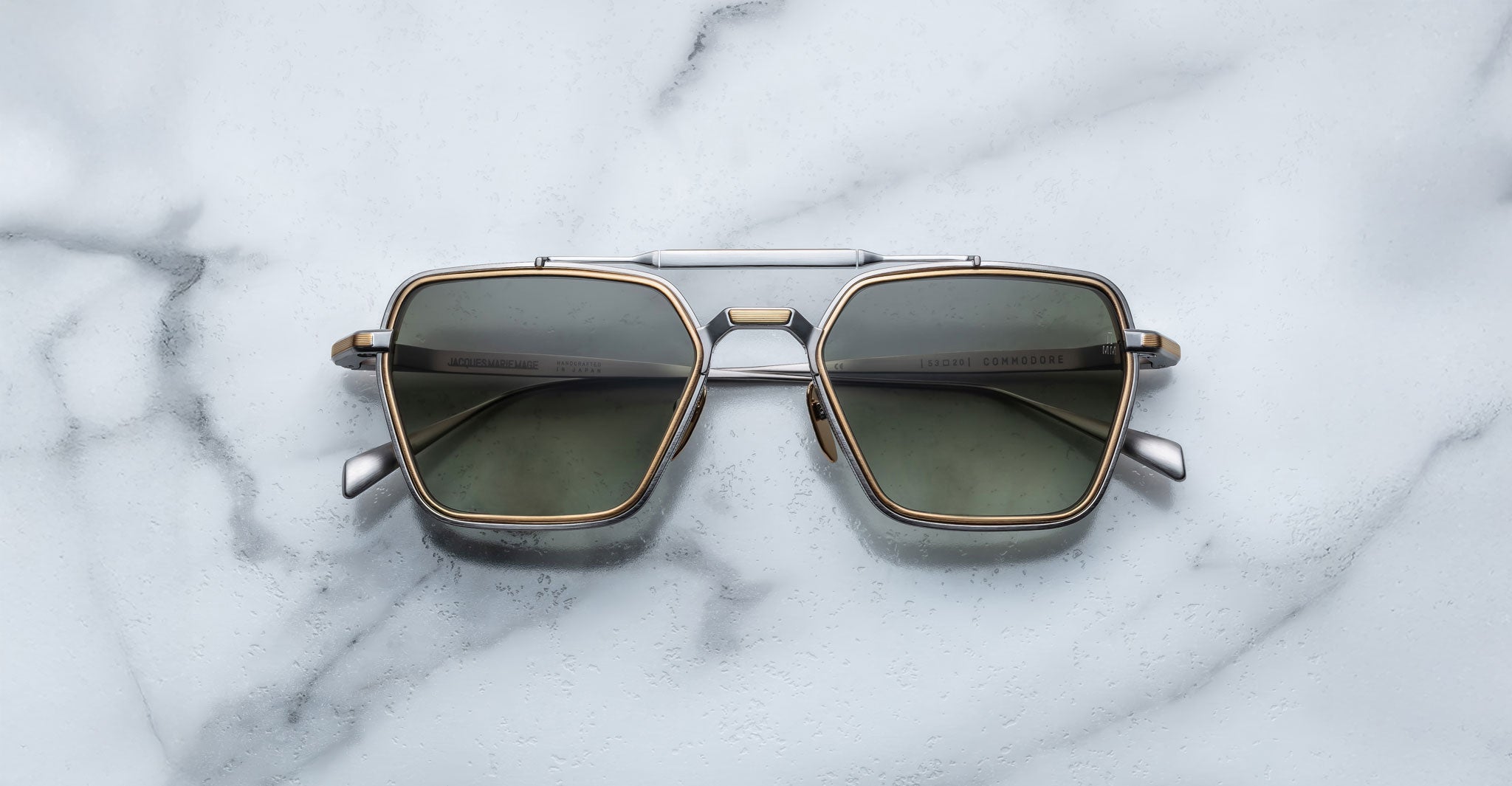 The Jacques Marie Mage Commodore sunglasses feature square metal frames with dark lenses and gold accents, shown resting on a white marble surface.