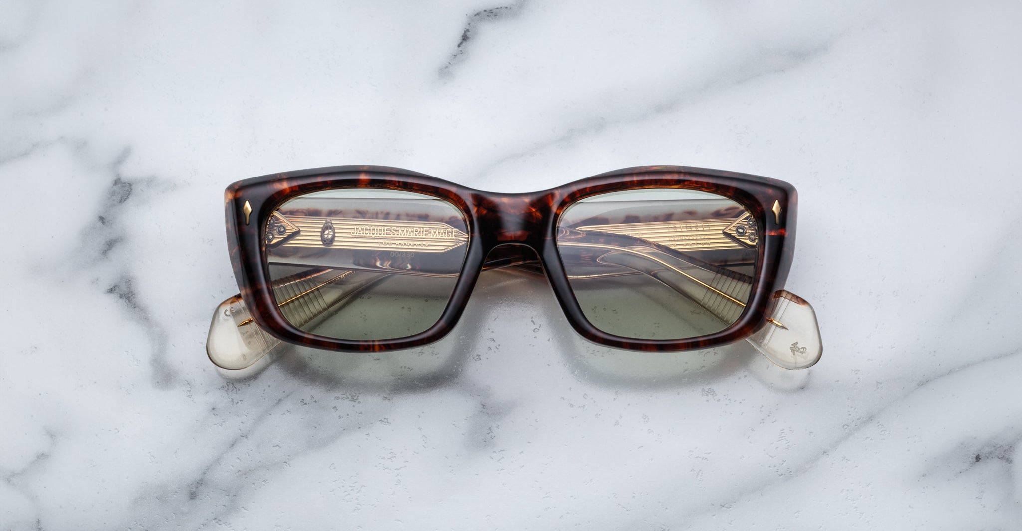 The Jacques Marie Mage Corniche sunglasses, featuring a rectangular tortoiseshell frame and slightly green-tinted lenses, rest on a white marble surface with the arms partially visible.