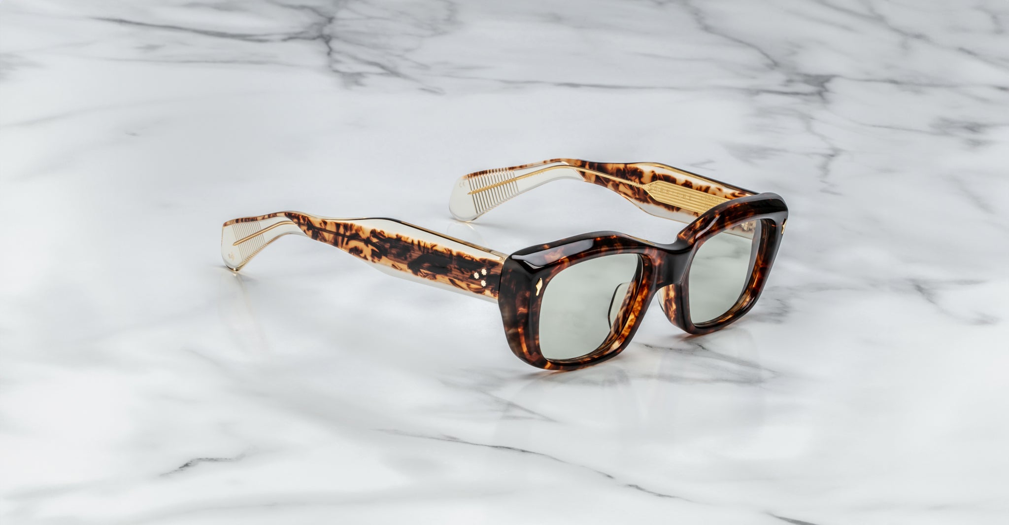 A pair of Jacques Marie Mage Corniche rectangular tortoiseshell sunglasses with thick frames and green-tinted lenses rests on a white marble surface.