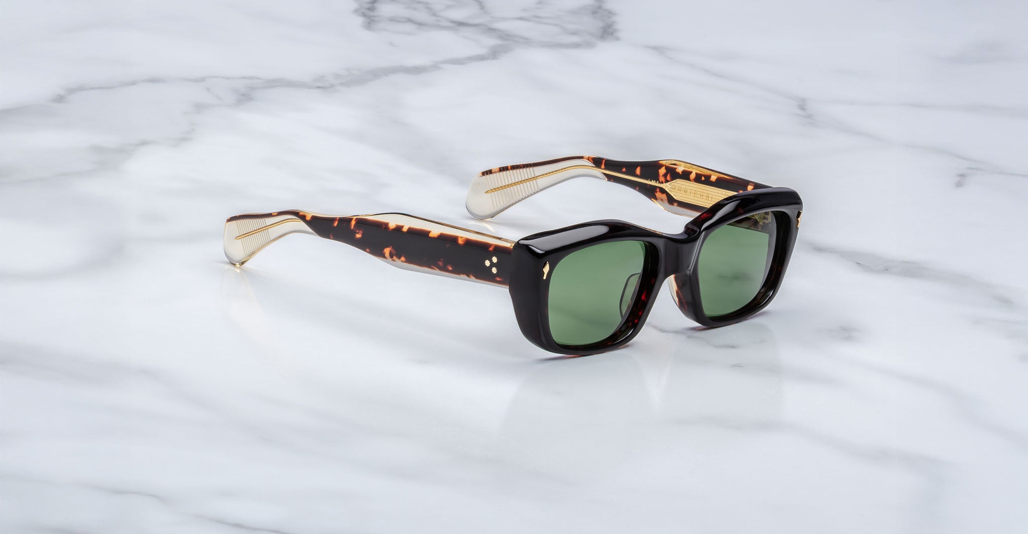 The Jacques Marie Mage Corniche sunglasses, featuring thick tortoiseshell frames and green-tinted lenses, rest on a white marble surface.