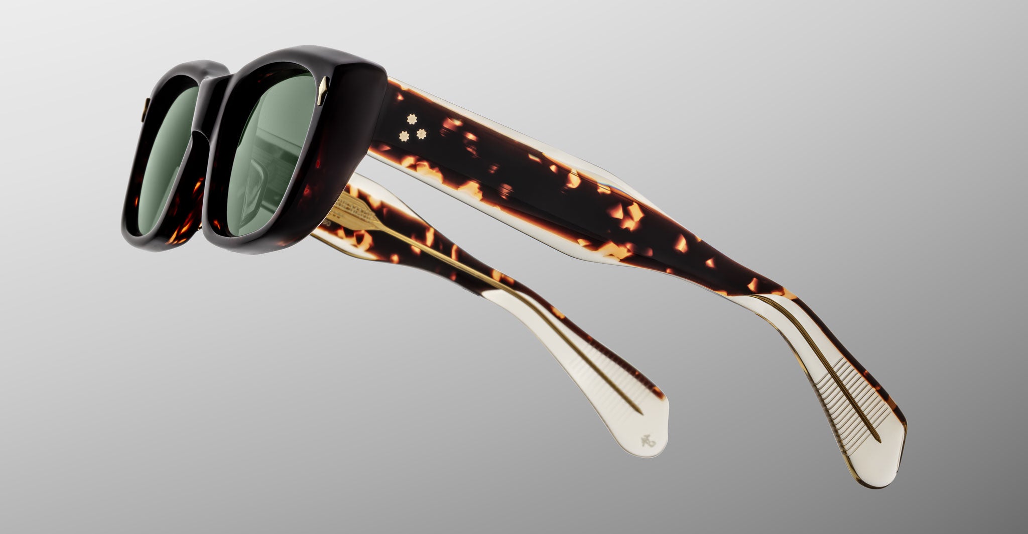 The Jacques Marie Mage Corniche sunglasses feature thick, square black frames, green lenses, and tortoiseshell-patterned temples with cream accents and engraved details, displayed on a light gray gradient background.