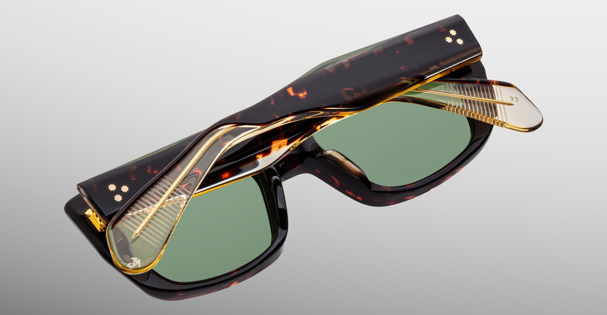 The Jacques Marie Mage Corniche sunglasses, featuring brown tortoiseshell frames, green lenses, and gold hinges, are folded neatly and displayed against a light gray background.