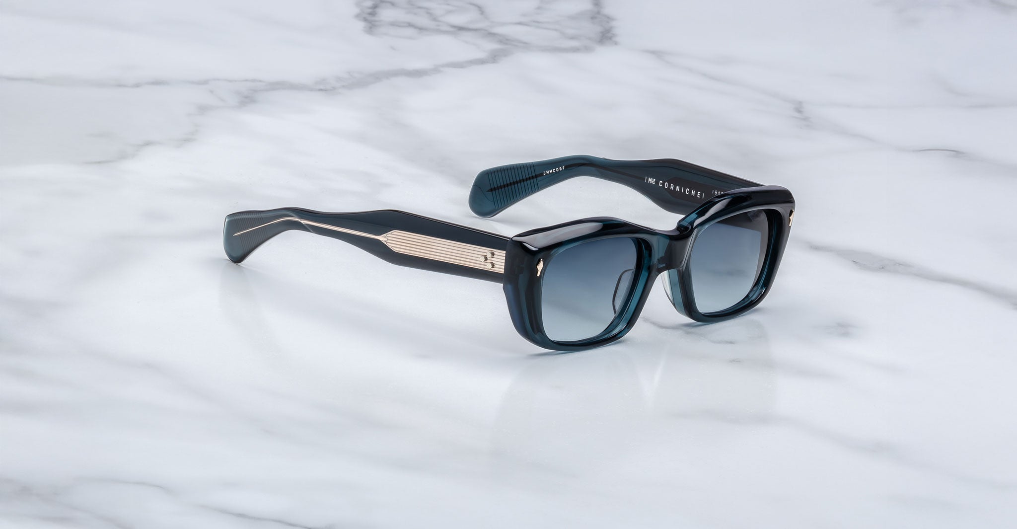 The Jacques Marie Mage Corniche sunglasses, with thick black frames, dark lenses, and metallic gold-accented temples featuring embossed branding near the hinges, rest on a white marble surface.