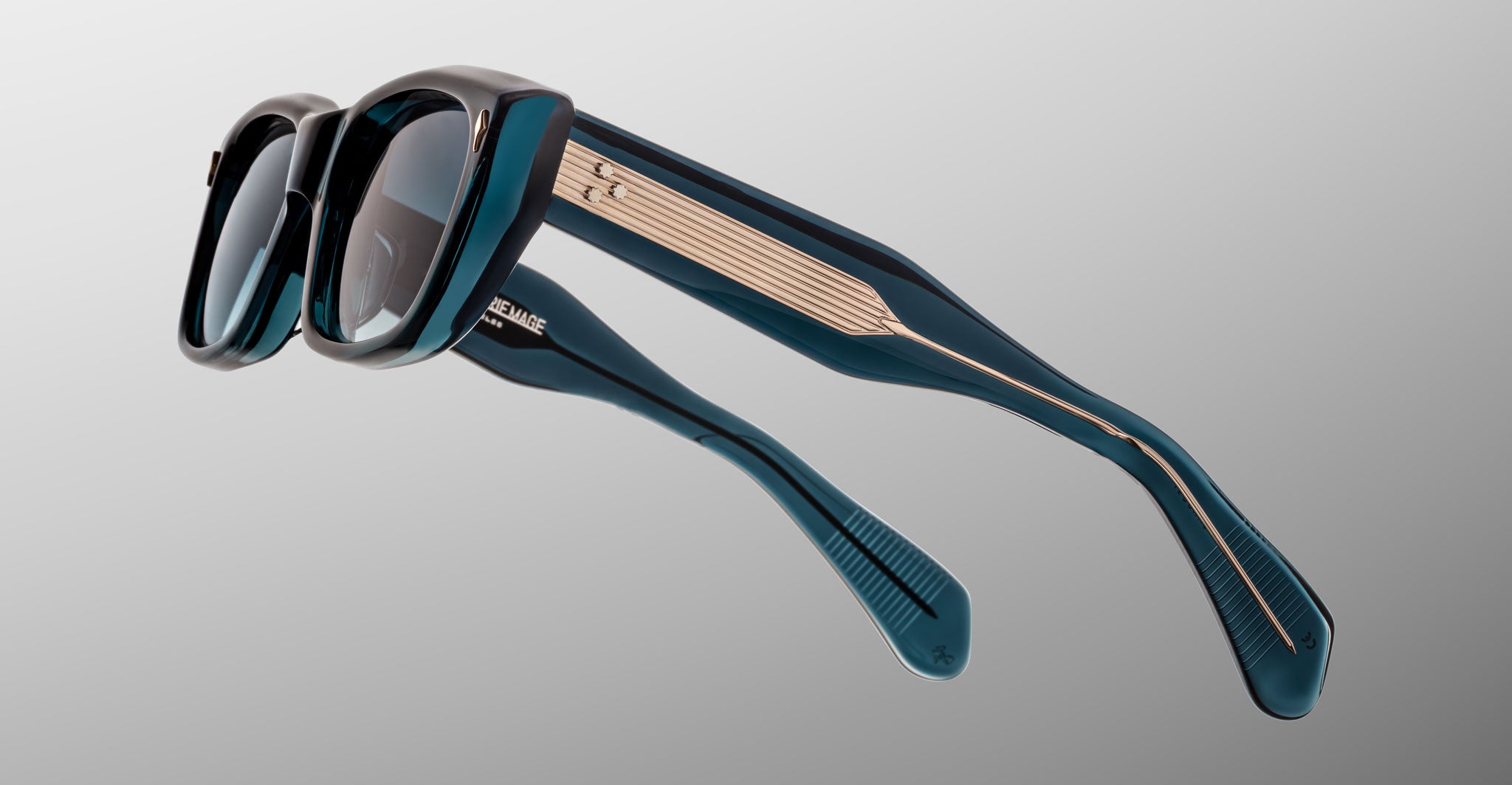 The Jacques Marie Mage Corniche sunglasses feature dark blue thick frames with gold detailing on the temples, displayed at an angle against a gradient gray background.