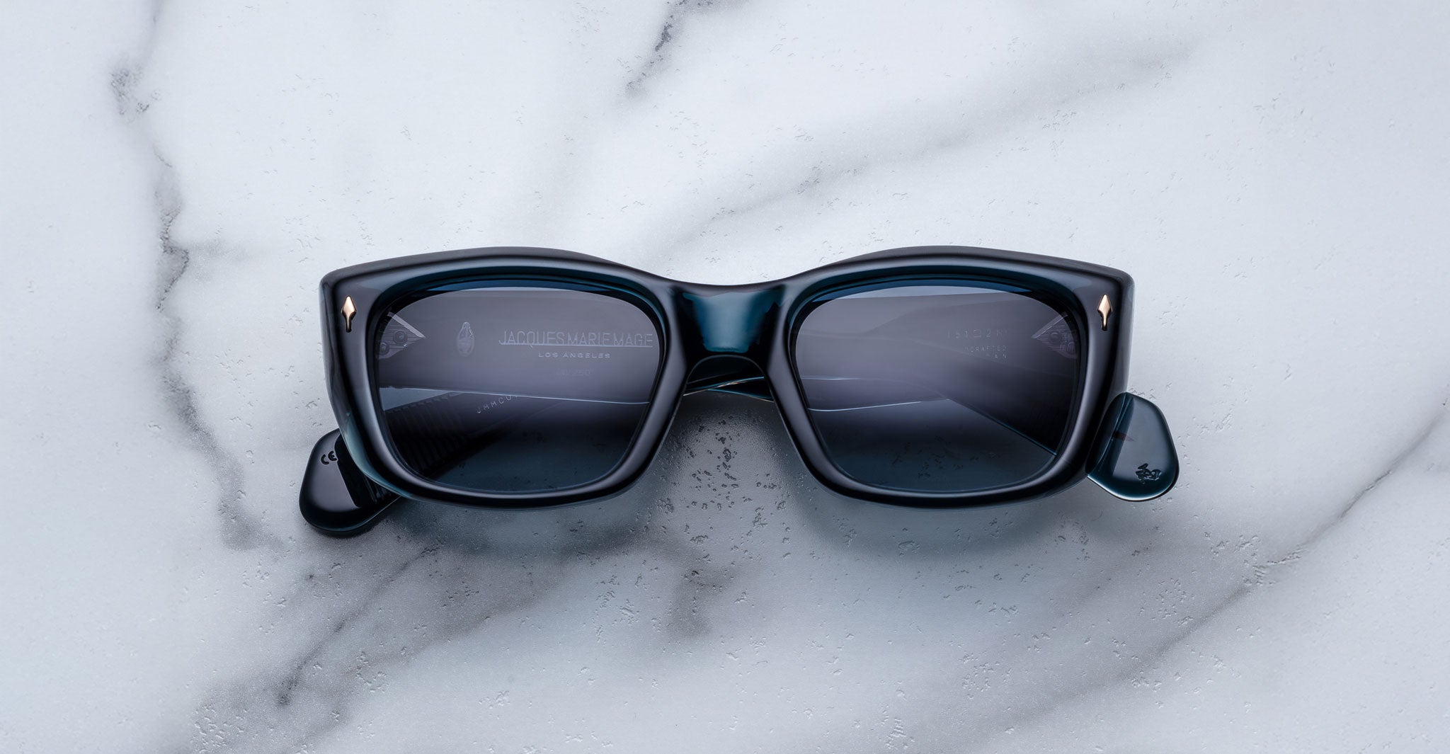 The Jacques Marie Mage Corniche sunglasses, featuring a black rectangular frame and dark lenses, rest on a white marble surface with gray veins. The arms of the sunglasses are folded.