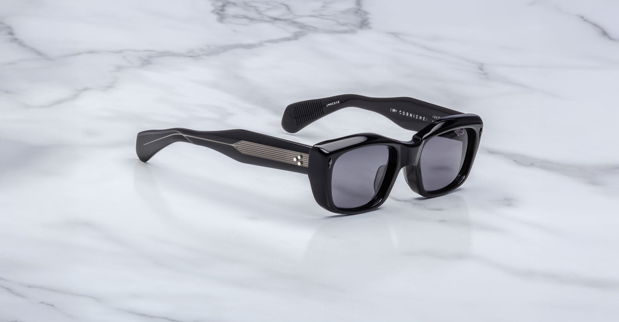 A pair of Jacques Marie Mage Corniche black rectangular sunglasses with thick frames and dark lenses rests on a white marble surface. The arms feature metallic accents with horizontal lines near the hinges.