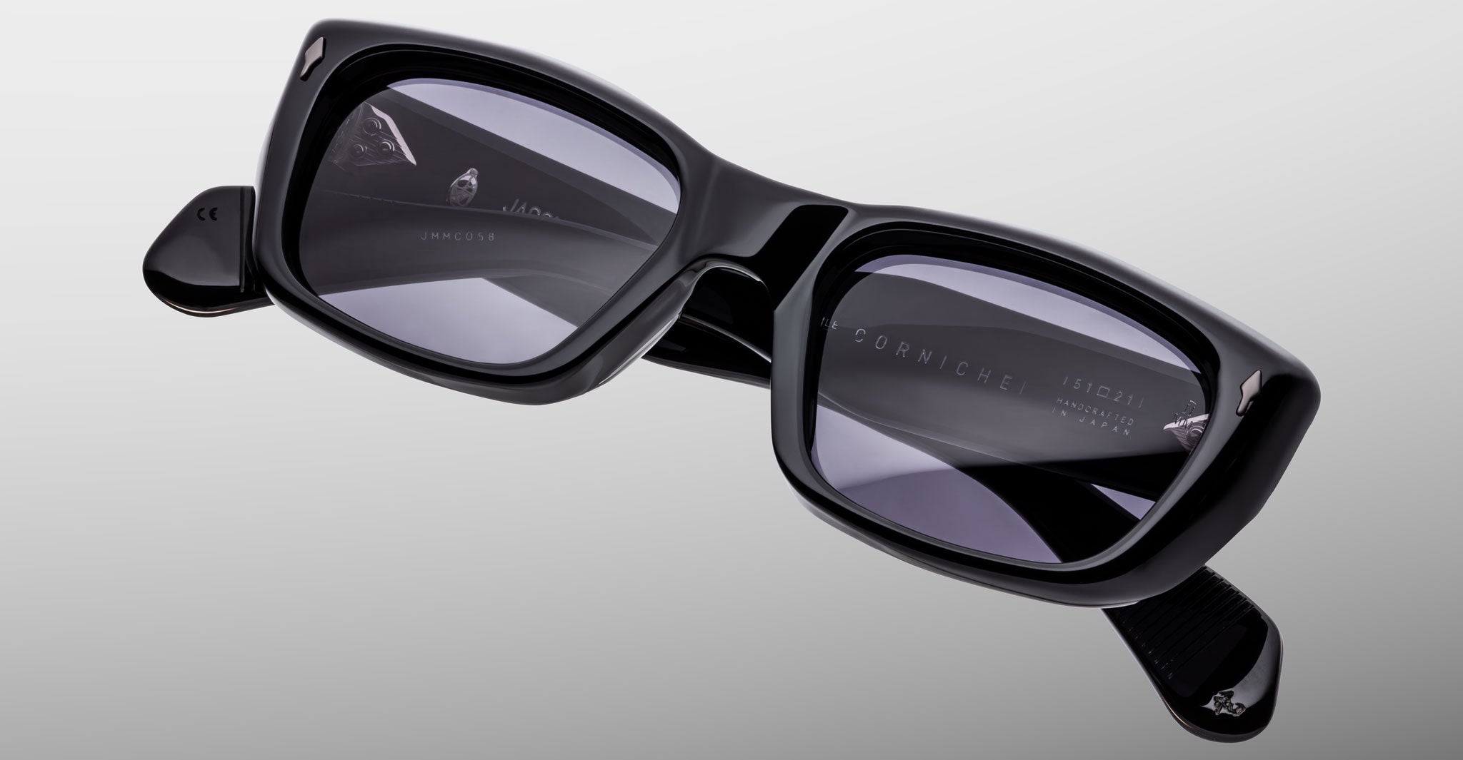 The Jacques Marie Mage Corniche sunglasses, featuring black thick-rimmed rectangular frames with dark lenses and folded arms, are displayed on a light gray background. Brand markings appear on the inside of the frames.