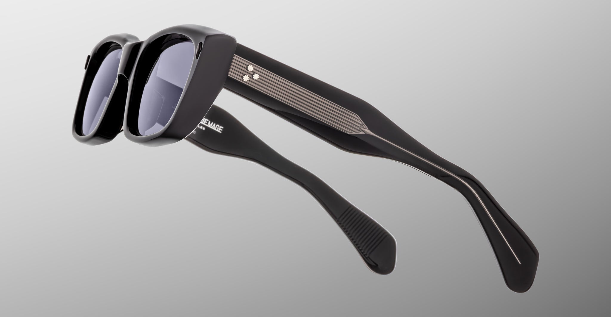 The Jacques Marie Mage Corniche sunglasses feature thick black frames and dark lenses, shown from a side and tilted angle on a plain gray background. The temples display metallic accent stripes and textured grips.