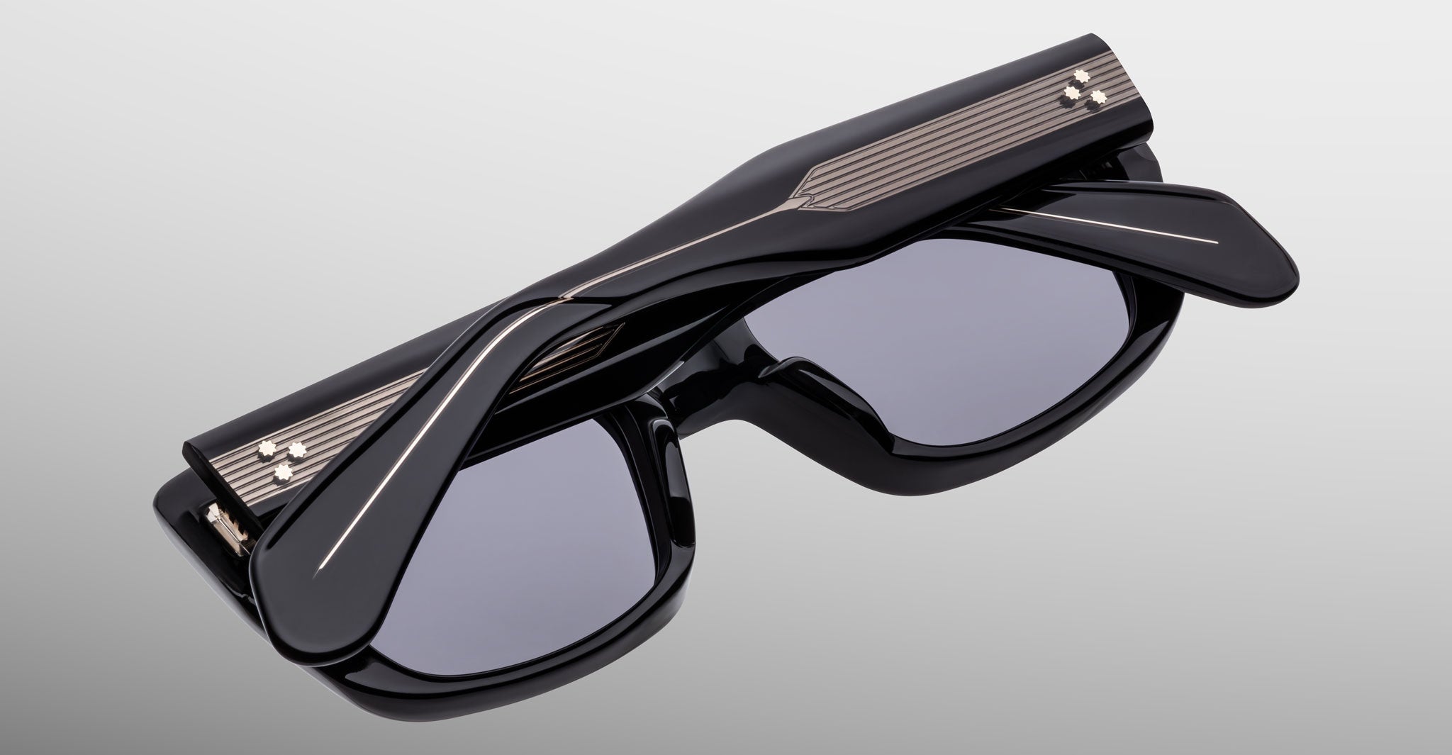 The Jacques Marie Mage Corniche sunglasses are shown folded, featuring thick black frames, dark lenses, metallic striped details on the temples, and small decorative studs, all set against a light grey background.