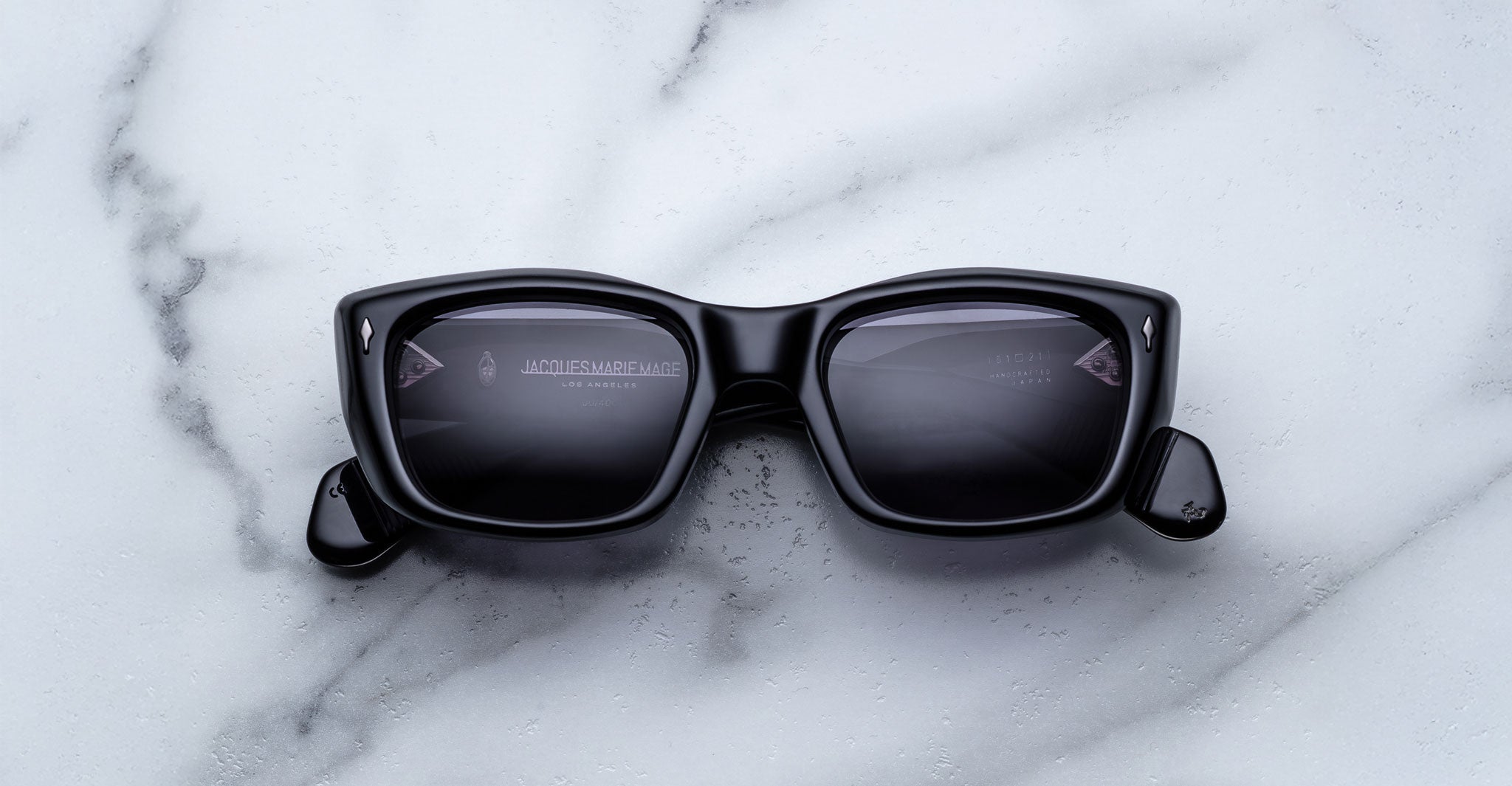 A pair of Jacques Marie Mage Corniche black rectangular sunglasses with dark lenses rests on a white marble surface. The arms are partially open, revealing faint text on the inside of the lenses and frame.