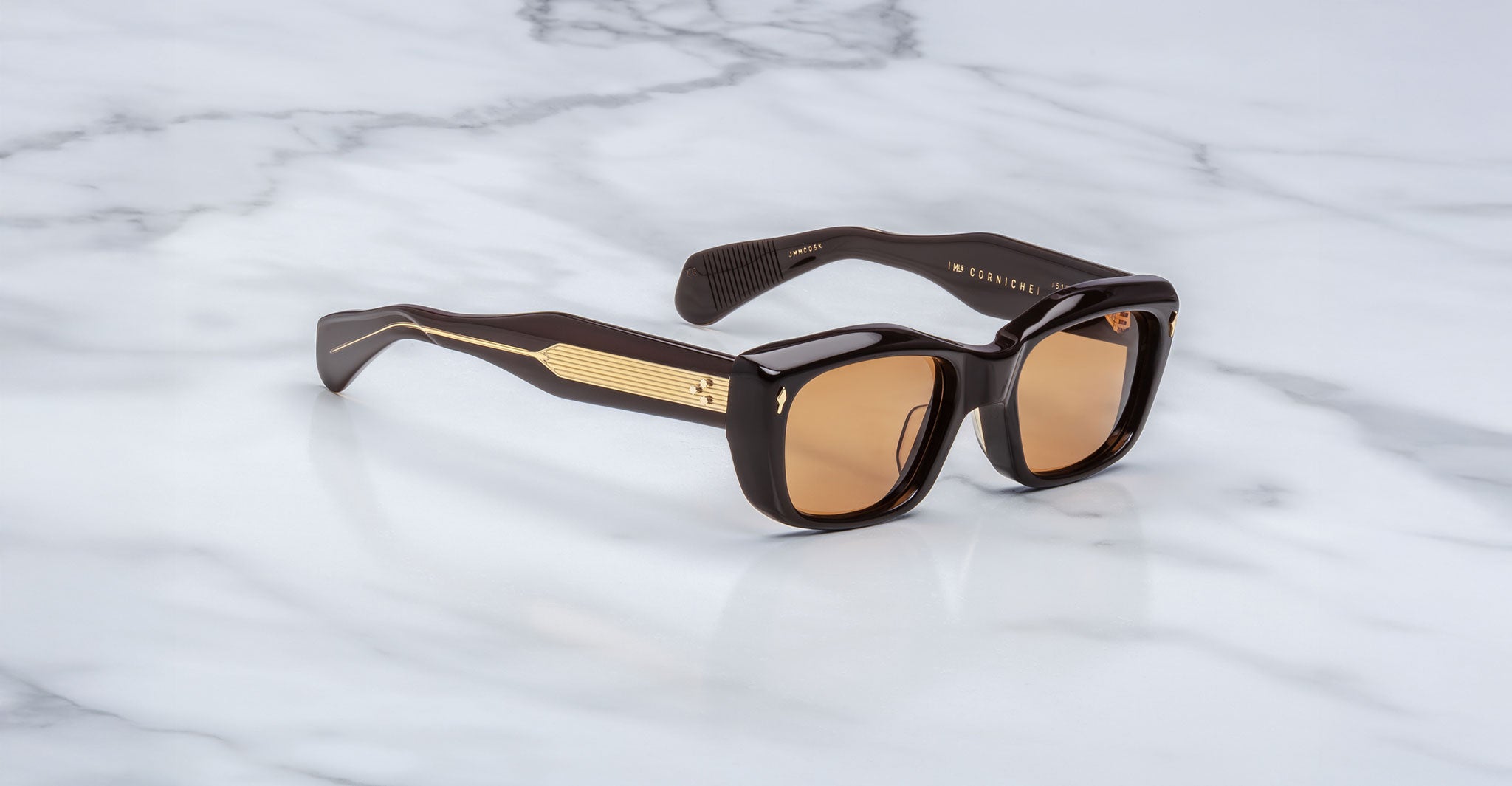 The Jacques Marie Mage Corniche sunglasses feature thick black frames, gold temple detailing, and brown tinted lenses, displayed on a white marble surface.