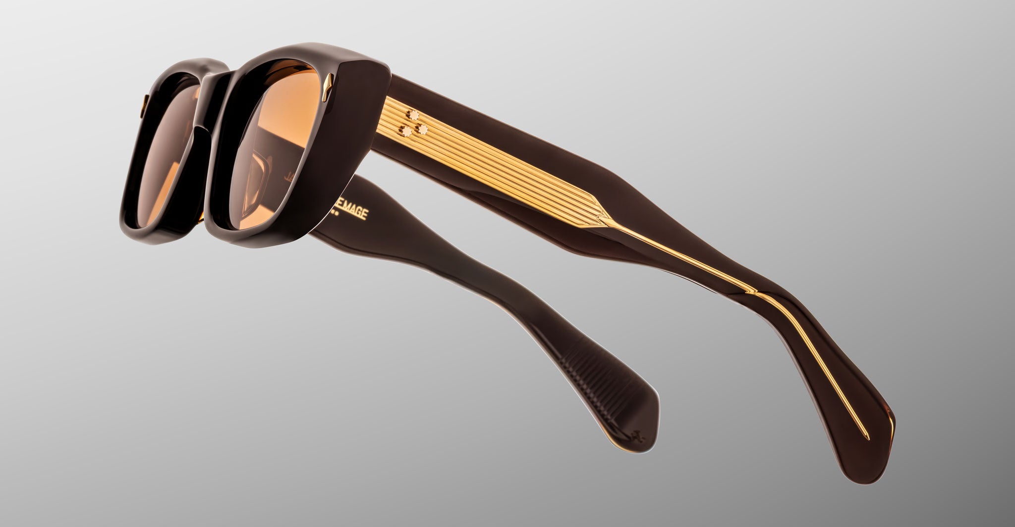 Close-up of Jacques Marie Mage Corniche sunglasses featuring dark brown, thick frames, gold detailing along the arms, and brown-tinted lenses, set against a gradient gray background.