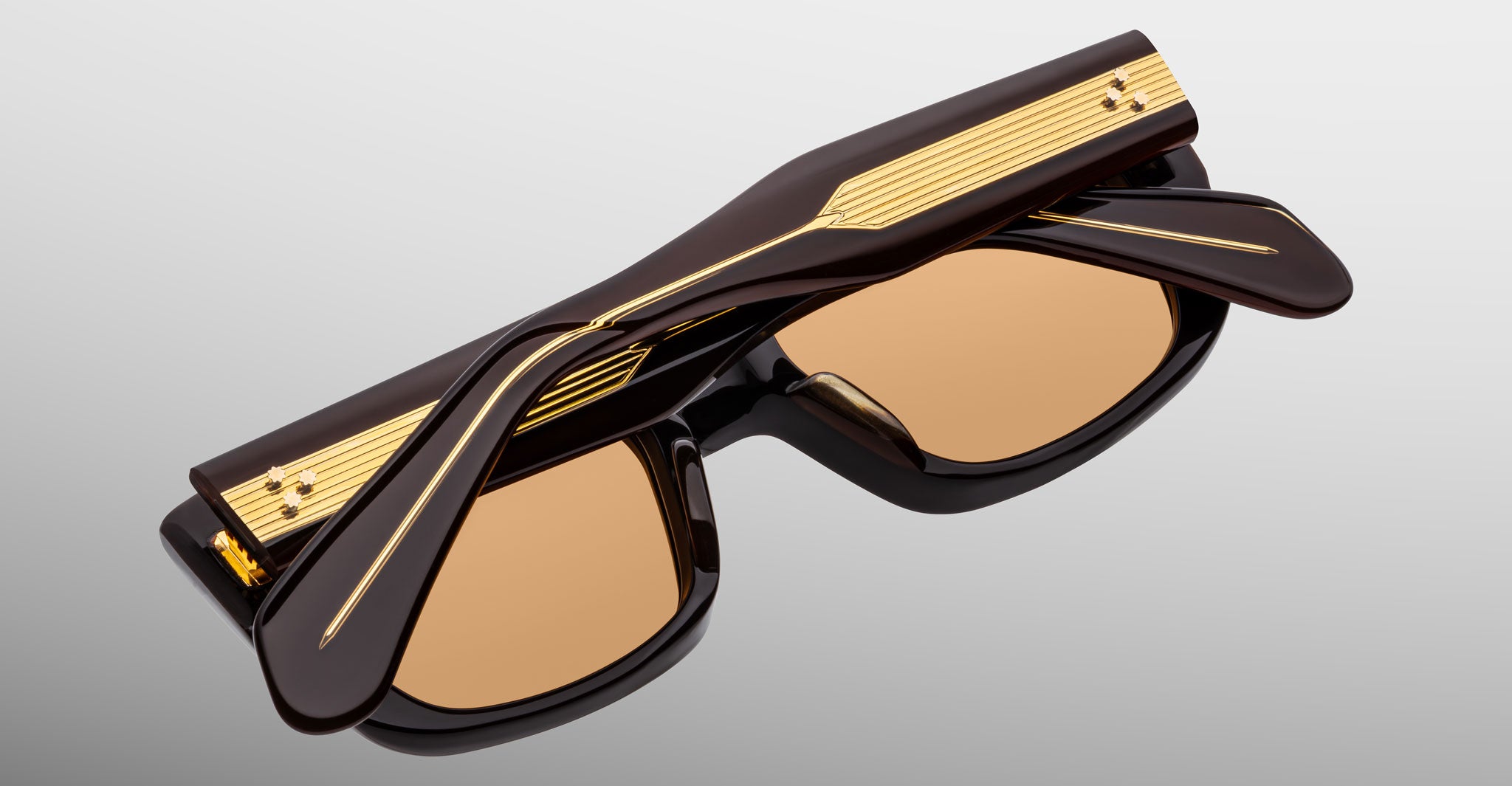 The Jacques Marie Mage Corniche is a pair of black sunglasses with brown-tinted lenses, gold accents, and decorative gold lines on thick arms, shown folded closed on a light gray background.