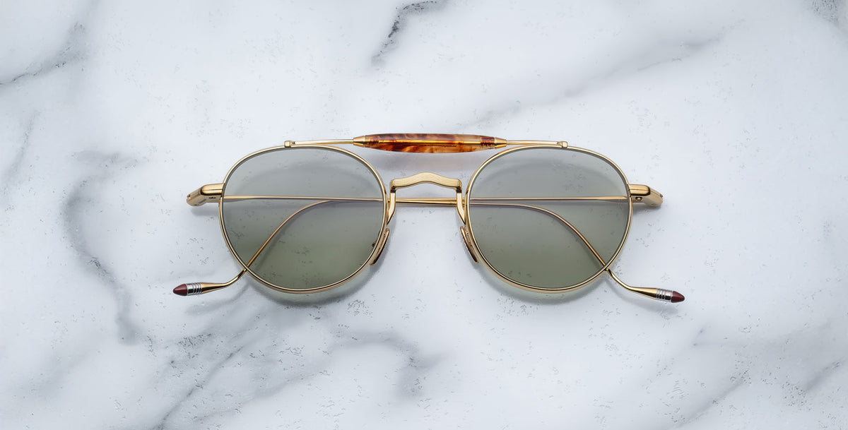 Front view of the Jacques Marie Mage Dasan titanium sunglass in gold color with light grey lenses on a white marbled with grey veining background