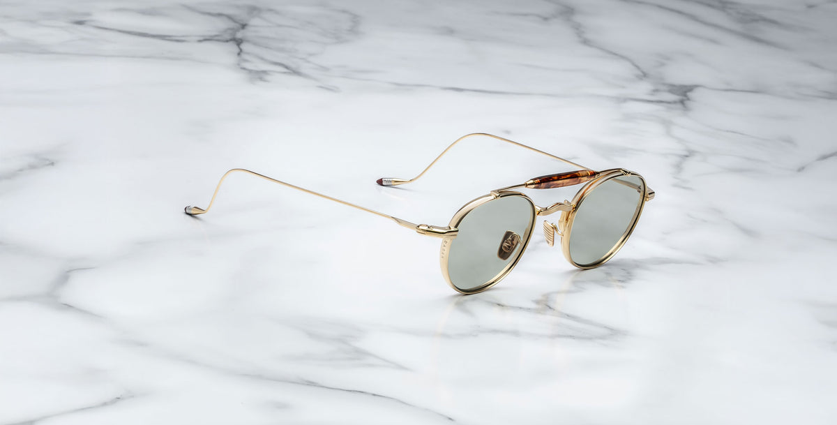 Profile view of the Jacques Marie Mage Dasan titanium sunglass in gold color with light grey lenses on a white marbled with grey veining background