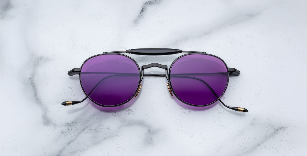 Front view of the Jacques Marie Mage Dasan titanium sunglass in black color with purple lenses on a white marbled with grey veining background