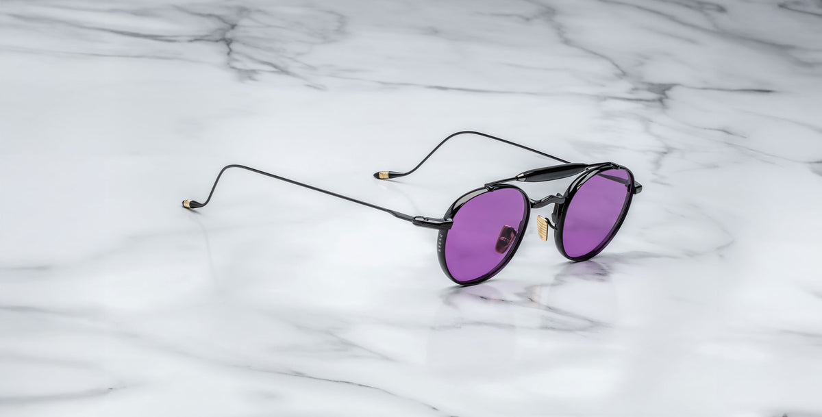 Profile view of the Jacques Marie Mage Dasan titanium sunglass in black color with purple lenses on a white marbled with grey veining background