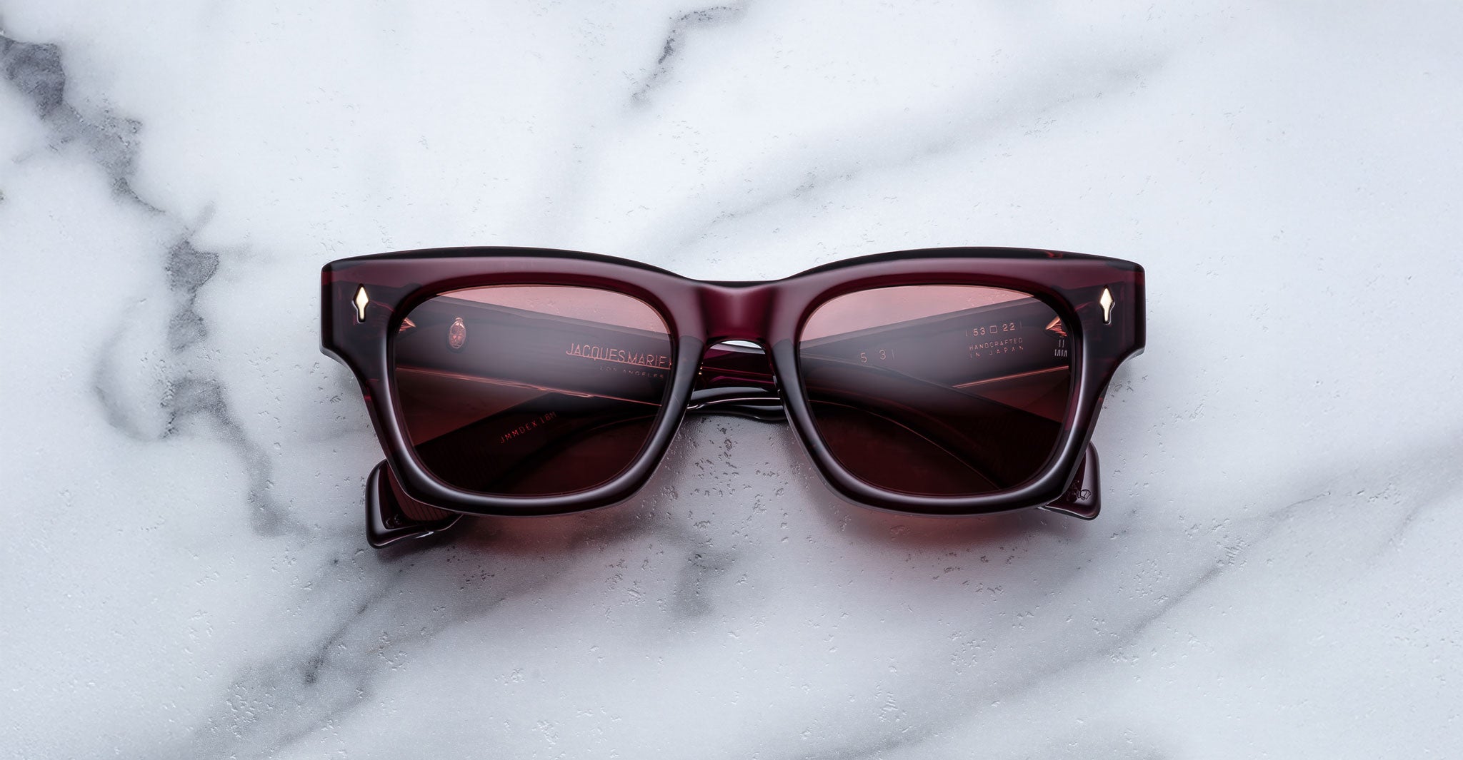 The Jacques Marie Mage Dealan 53 sunglasses, featuring a rectangular burgundy frame and dark lenses, rest on a white marble surface. Gold text and subtle decorative details accent the inside of the arms.