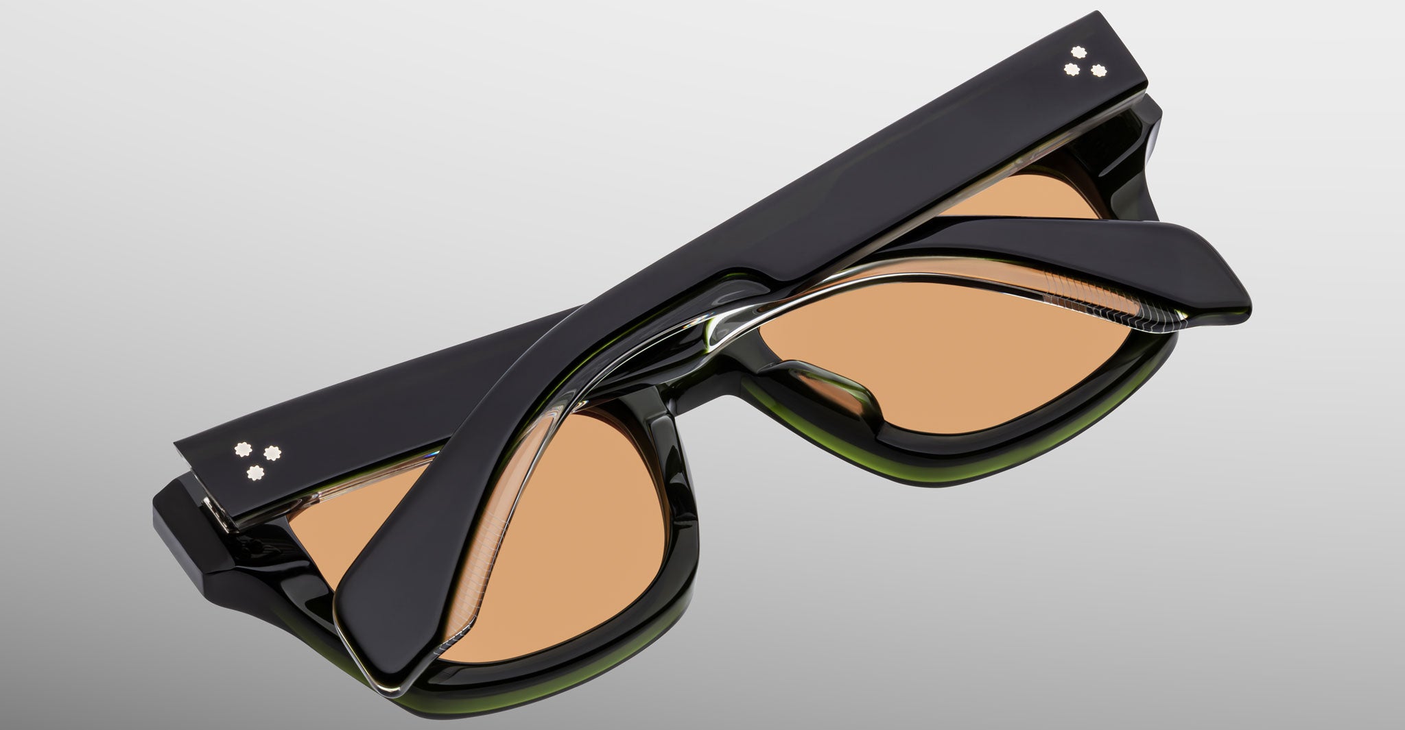 The Jacques Marie Mage Dealan 53, a pair of black sunglasses with thick frames and peach-tinted lenses, is folded and viewed from above on a light gray background, showing three small dots on the temples near the hinges.